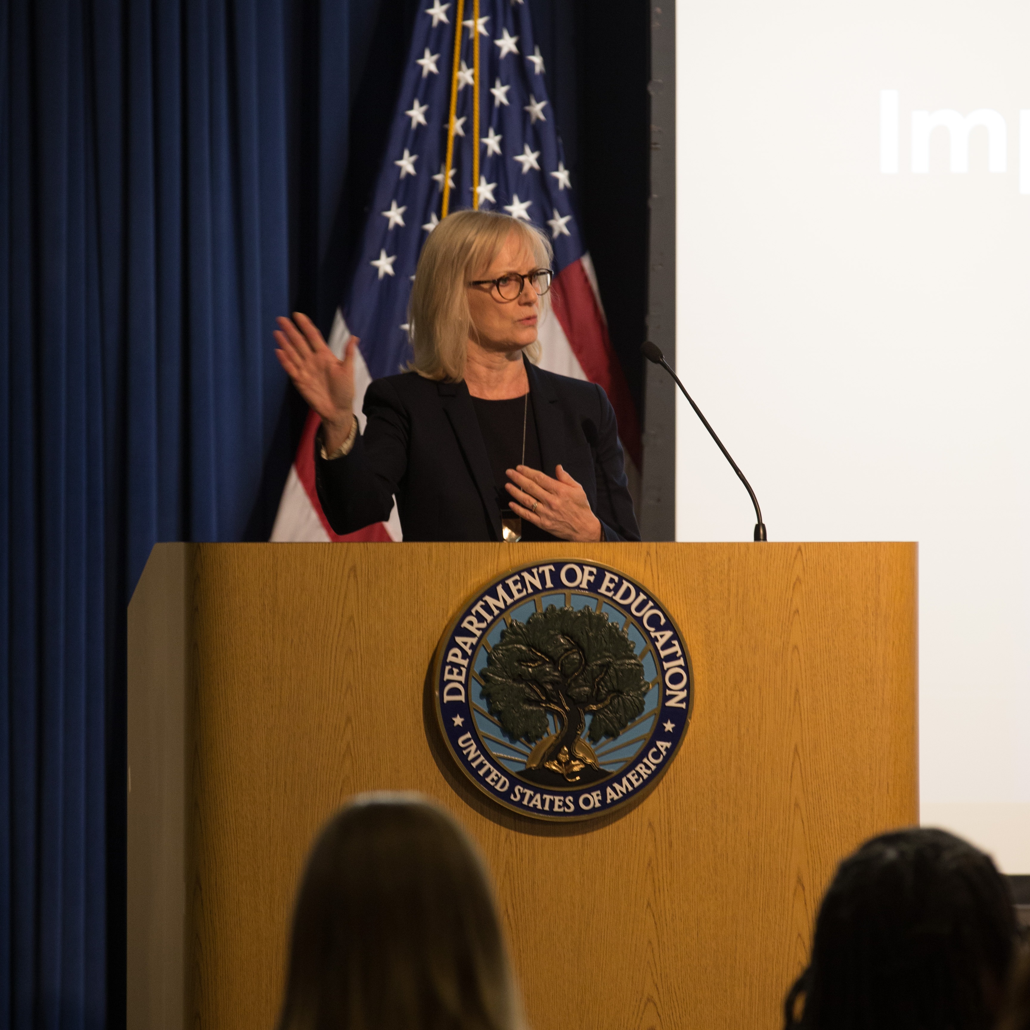 Woman speaking at podium in front of an American flag, Department of Education seal, and audience.