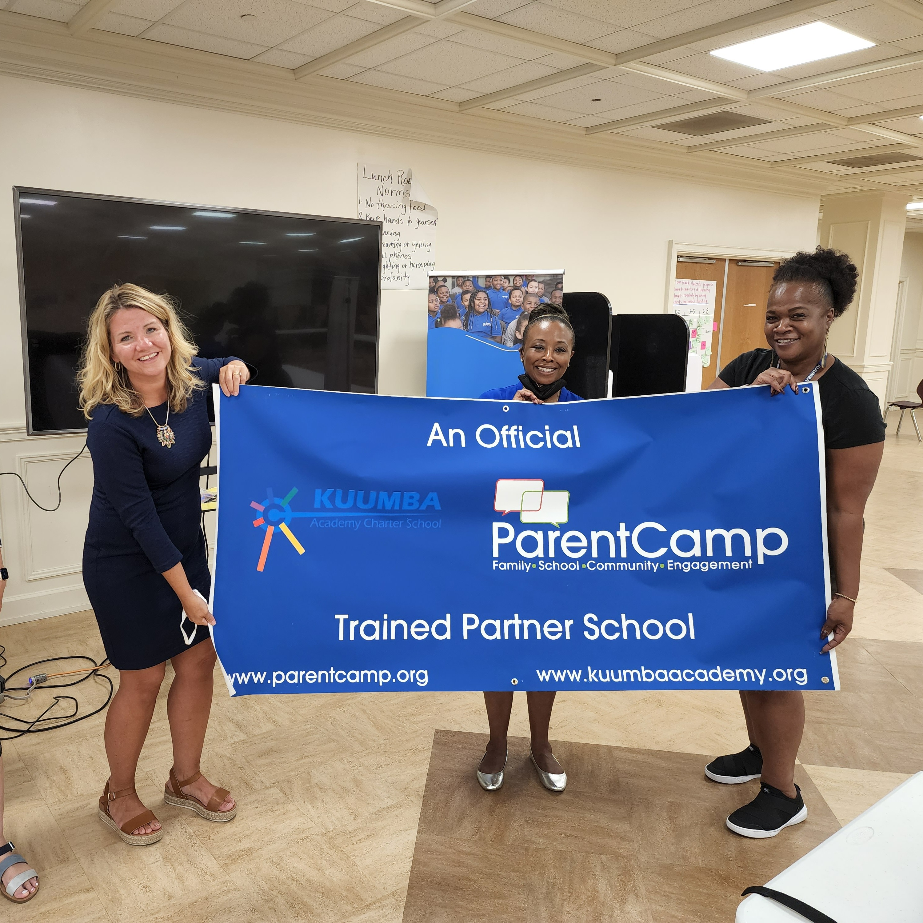 Four adults in blue shirts hold a blue banner that reads "ParentCamp". They stand in a room with a TV and chairs.