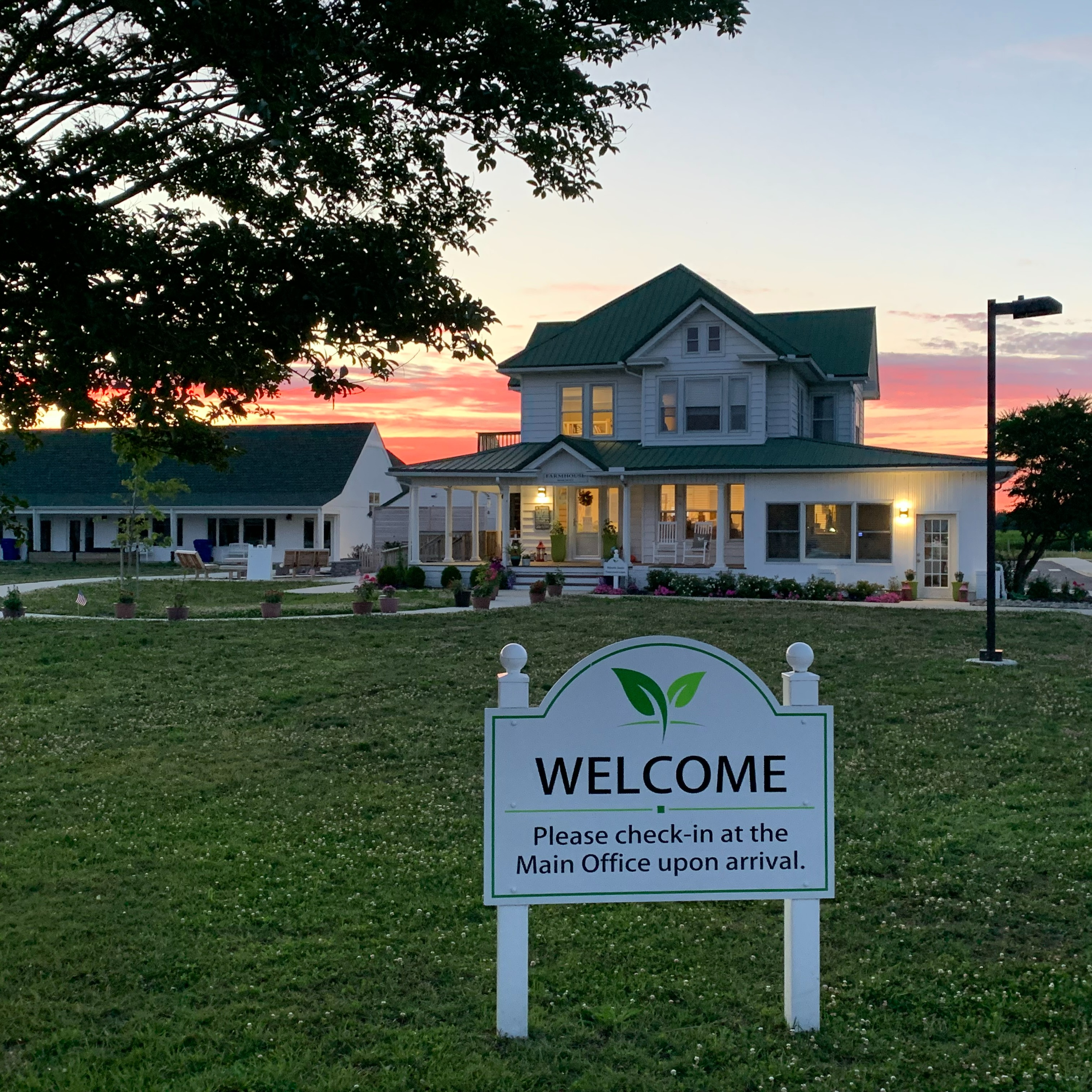 A welcoming sign in a grassy area in front of a large building. The background shows sunset colors and a clear sky.