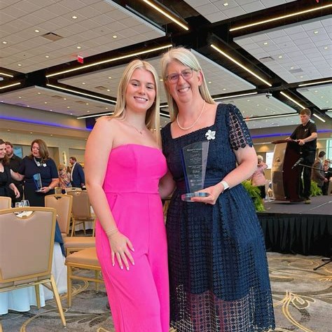 Two women at an event, one smiling, the other holding an award. Behind them is a stage with a person.