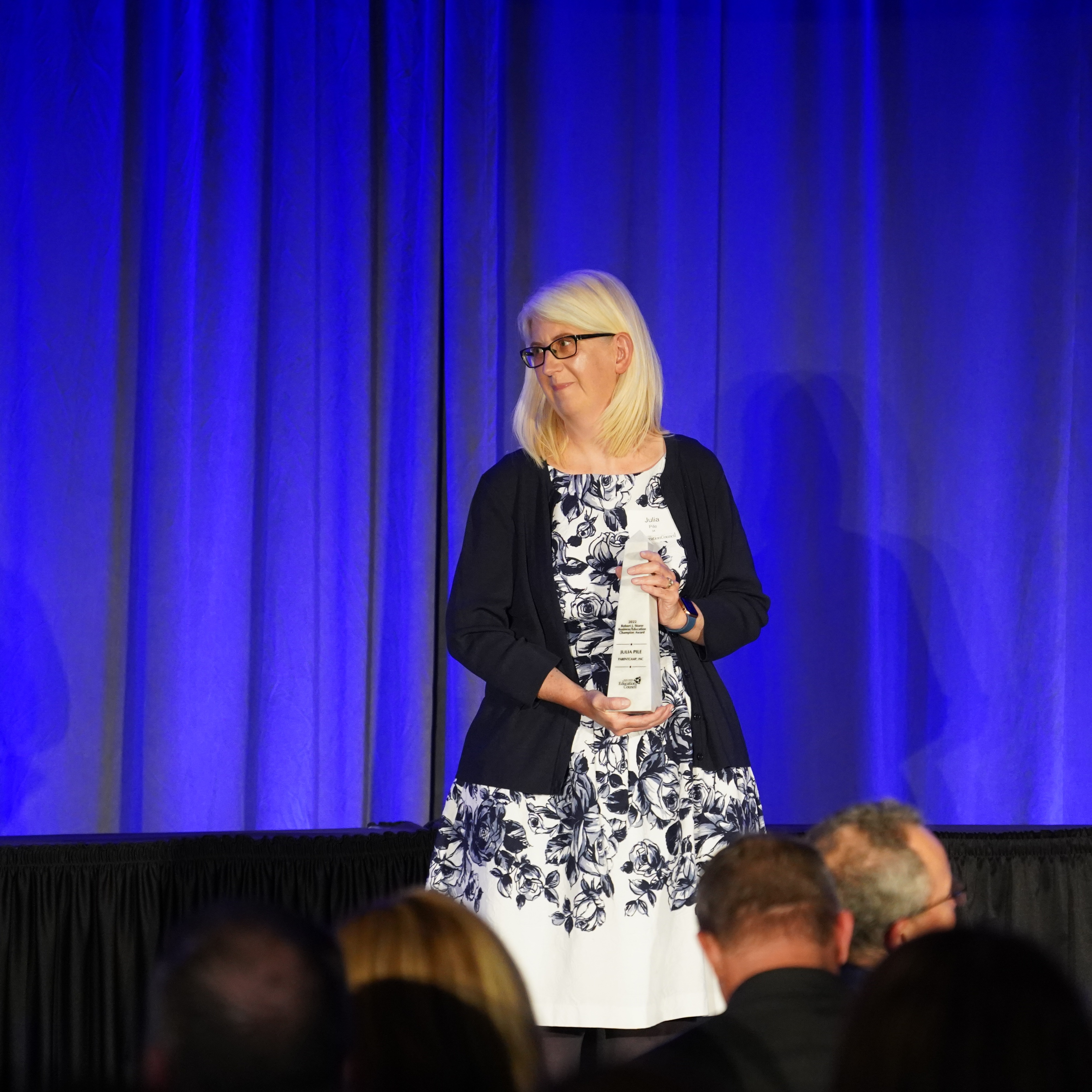 A woman in a floral dress stands on stage holding an award, with blue curtains behind her.