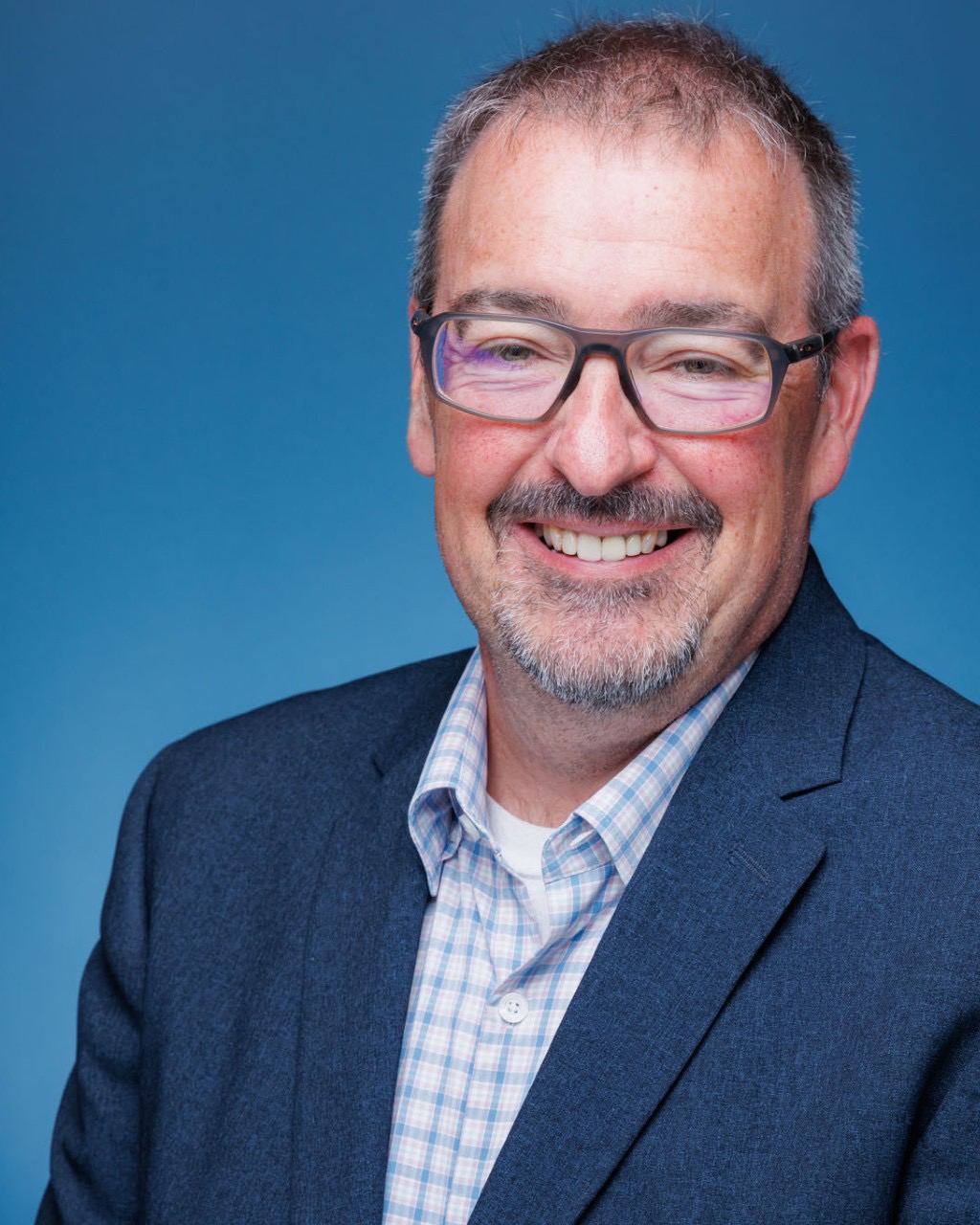 A man in glasses smiles against a blue backdrop, wearing a suit and plaid shirt, with short hair.