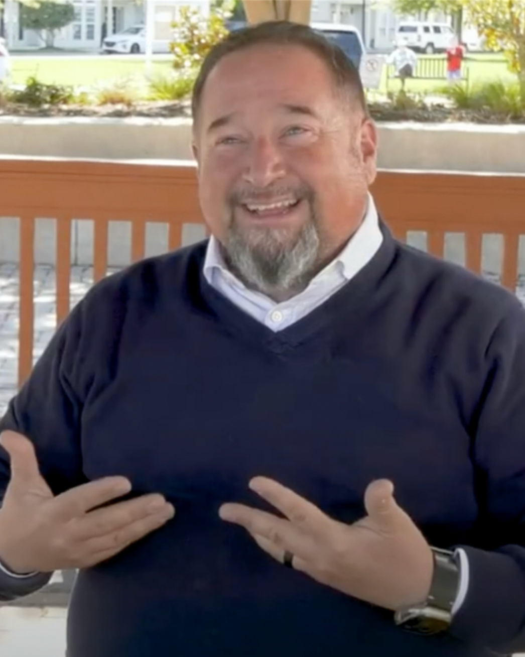 A person in a blue sweater smiles and gestures with hands on a porch with a wooden railing.