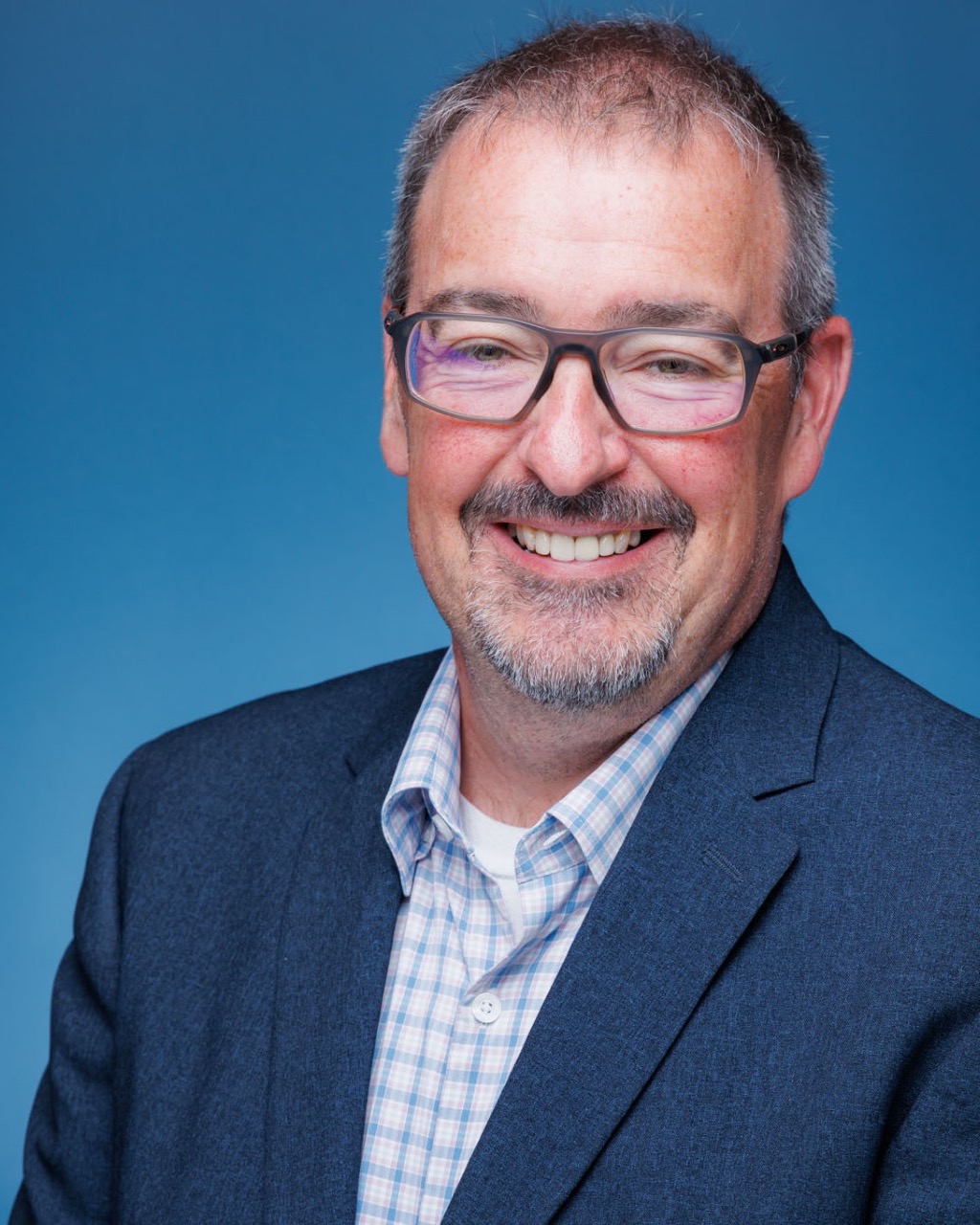 A man with a beard and glasses, wearing a blue coat, white shirt, and checkered shirt underneath, smiles against a blue background.