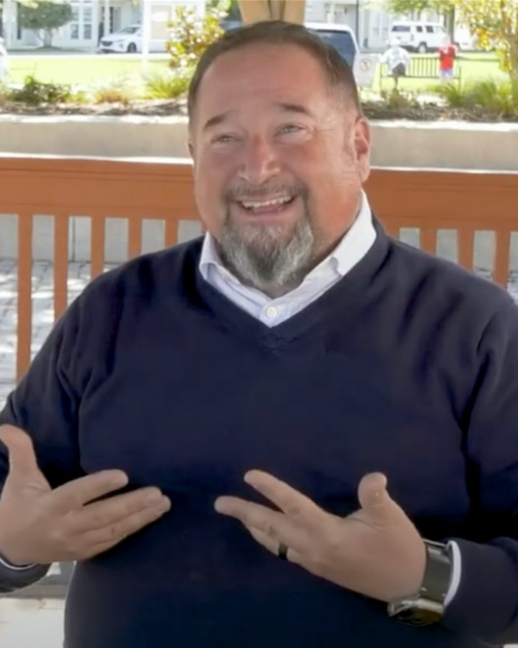 Man in a navy sweater with a white undershirt, gesturing with hands, smiling, standing on a porch.