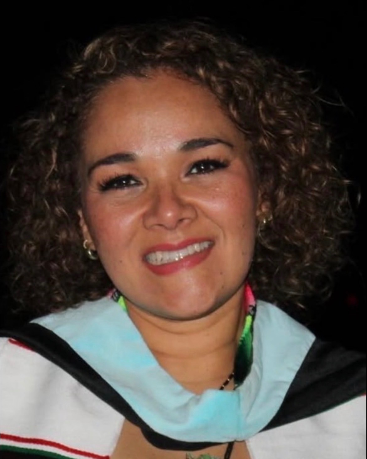 Woman with curly hair smiles at the camera. Wearing a graduation gown, scarf, and necklace.