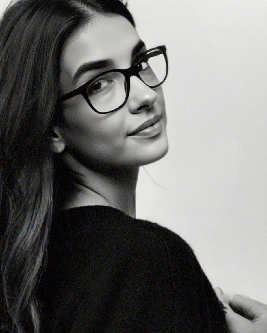 A black-and-white photo of a woman with long hair, wearing glasses, smiling, and touching her hair.