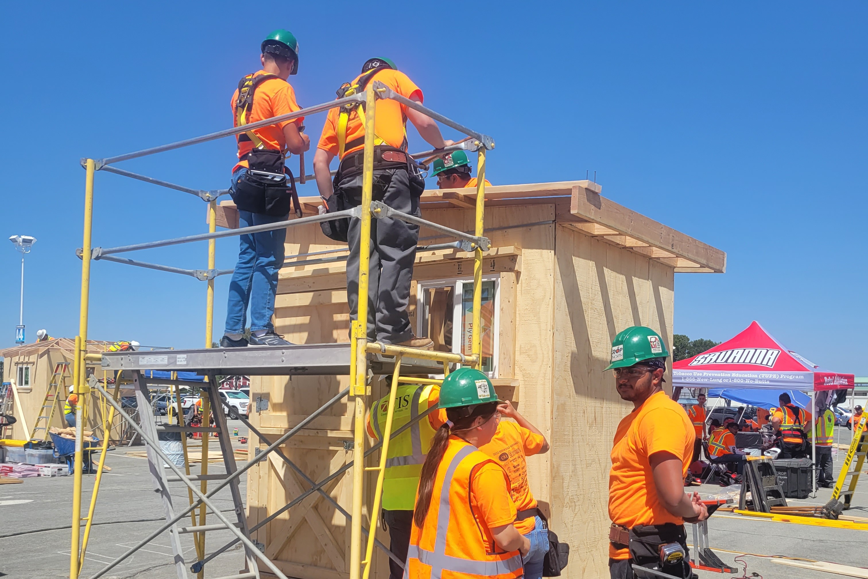 a group of construction workers work on a tiny house. two people are on a scafold and two others are talking near the tiny house