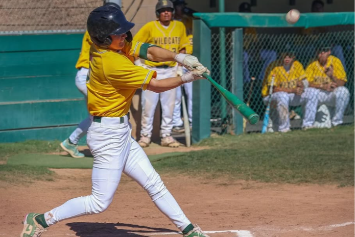 a baseball player dressed in white pants and yellow jersey swings at a baseball while the dugout can be seen behind him