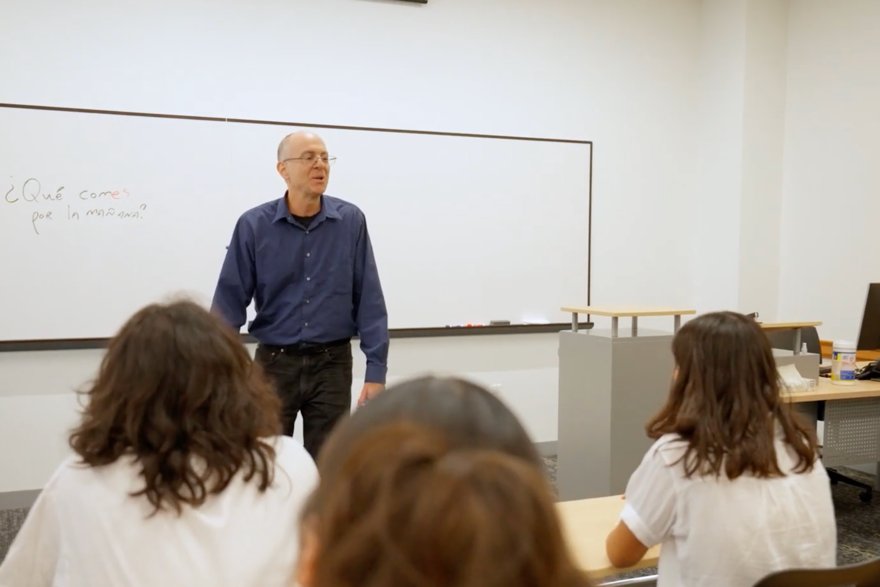 a professor at the front of a class standing in front of a whiteboard. You can see the backs of students heads