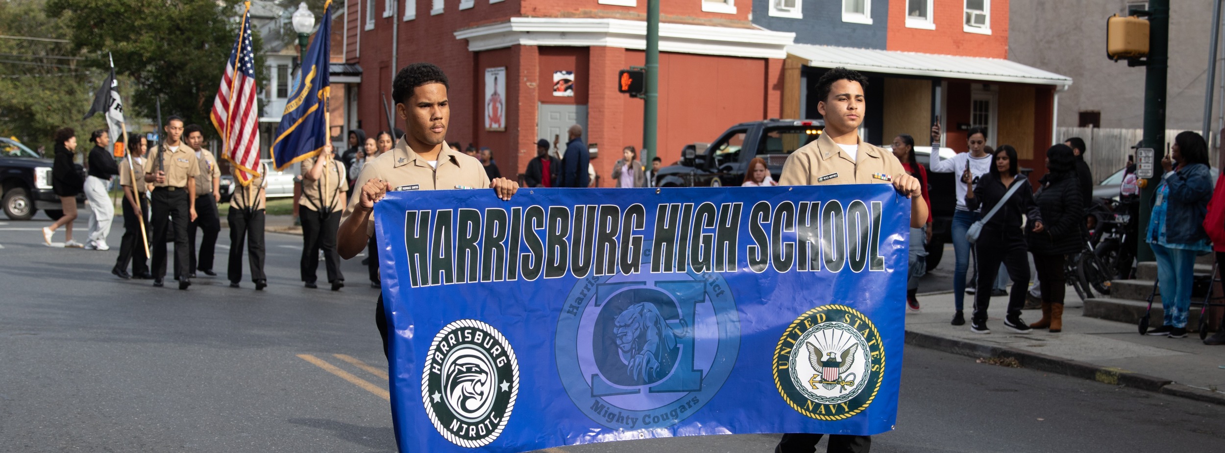 Two Harrisburg High School NJROTC cadets march in a community parade carrying a blue banner that reads “Harrisburg High School,” while other cadets with flags and spectators line the street behind them.