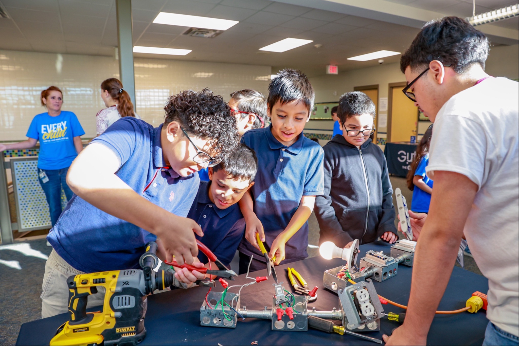 Several children work on electronic projects with tools. A man in a white shirt assists them. The area has a table and overhead lights.