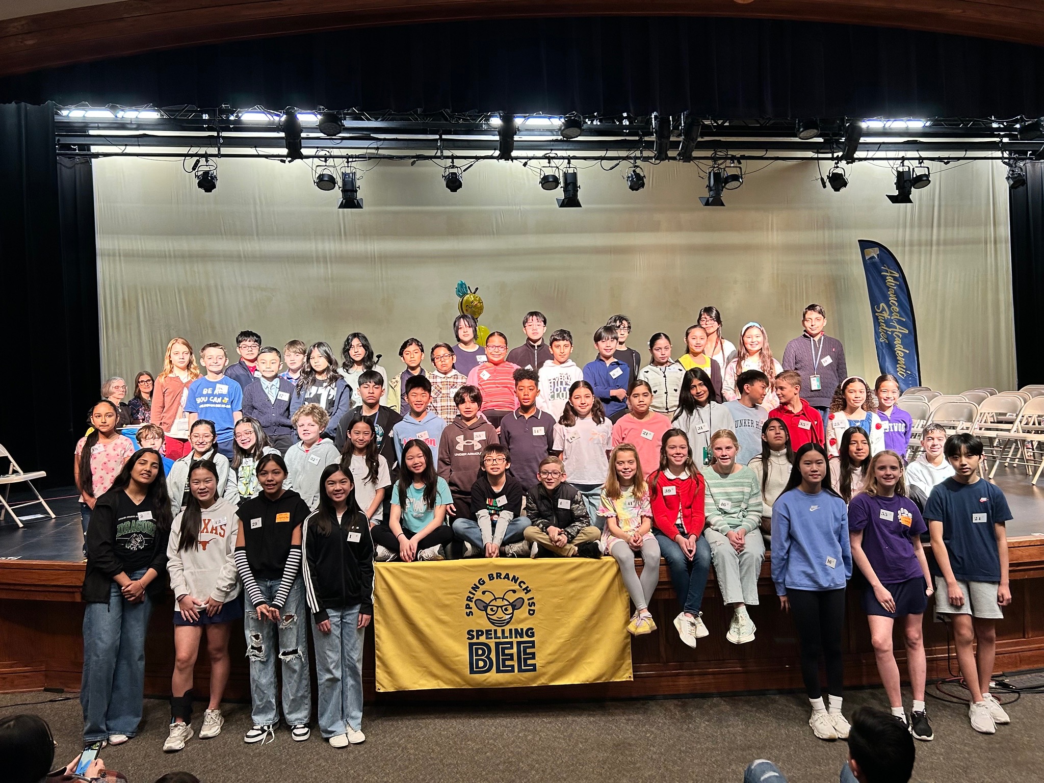 Group of students on stage, some sitting on the floor, others standing. Banner reading "SPELLING BEE" on front. Stage backdrop, chairs in background.