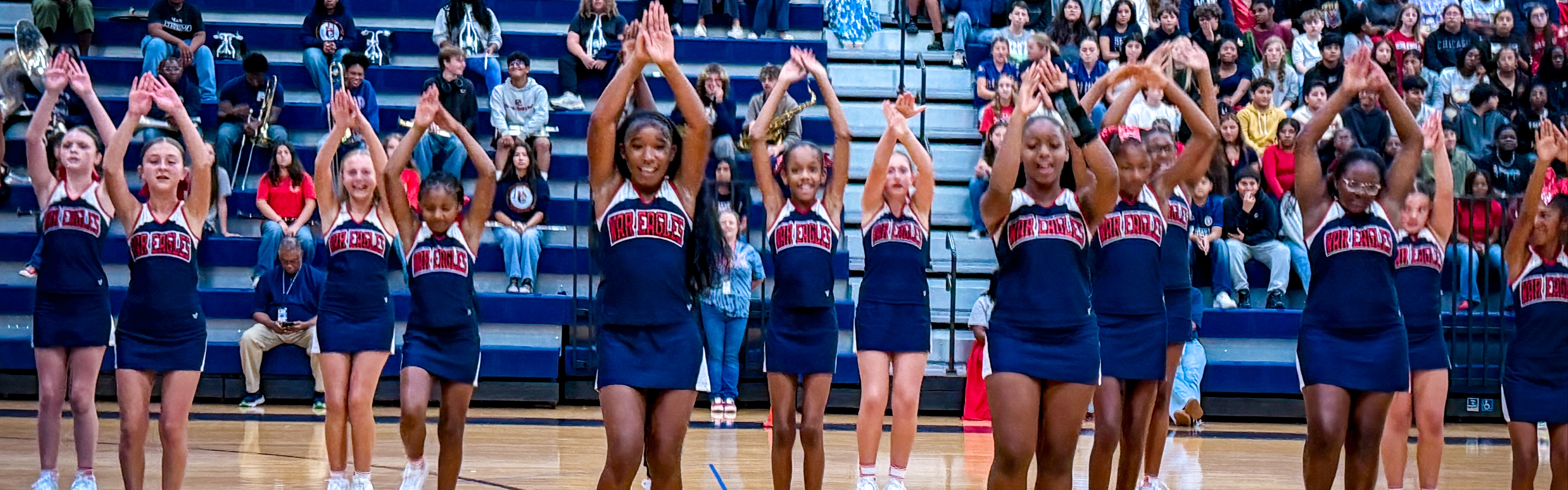 A cheerleading squad on a basketball court performs synchronized moves. They wear matching blue uniforms and white shoes. Spectators sit in bleachers.