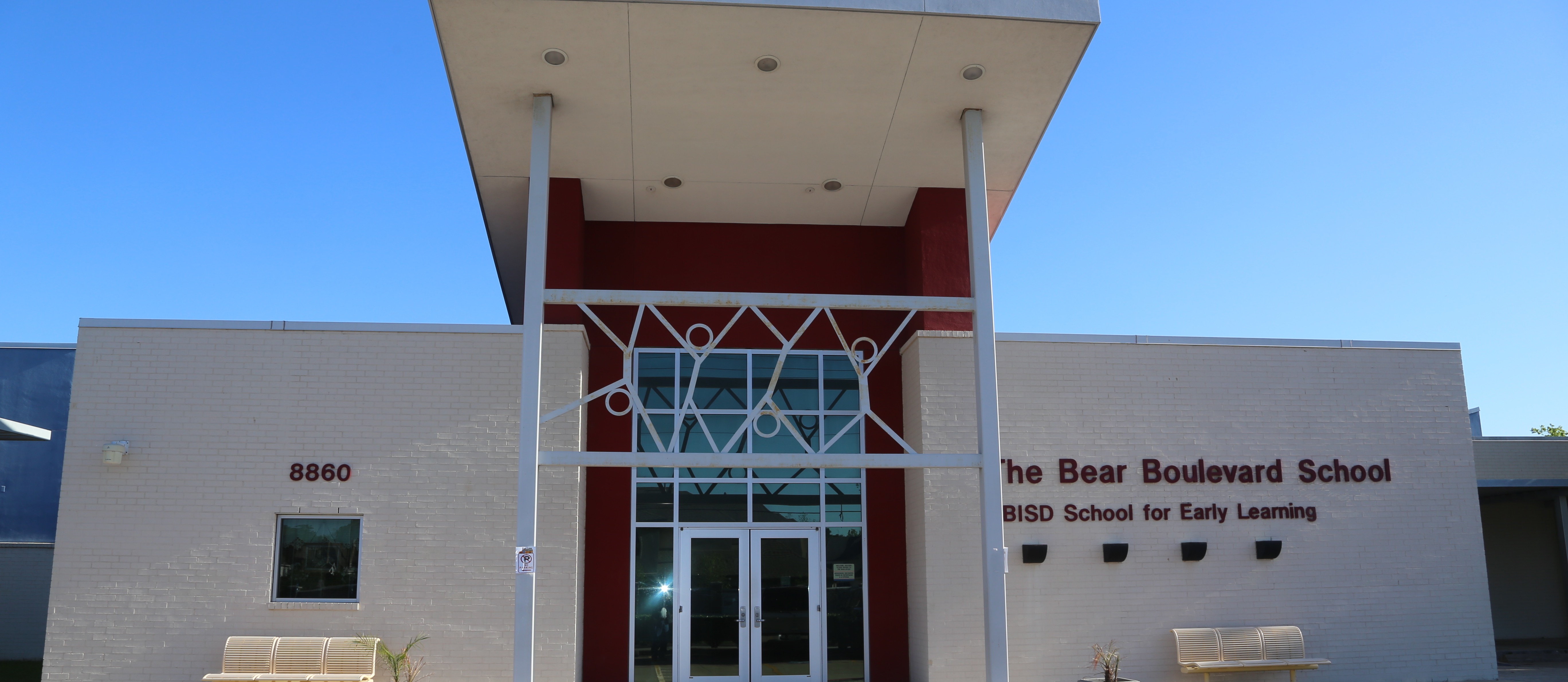 A white building with a red accent. Two benches and potted plants. The name reads "The Bear Boulevard School."