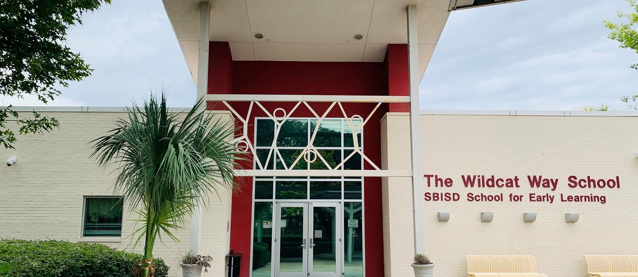 Entrance of The Wildest Way School, a white building with red accents, a glass door, and palm trees.
