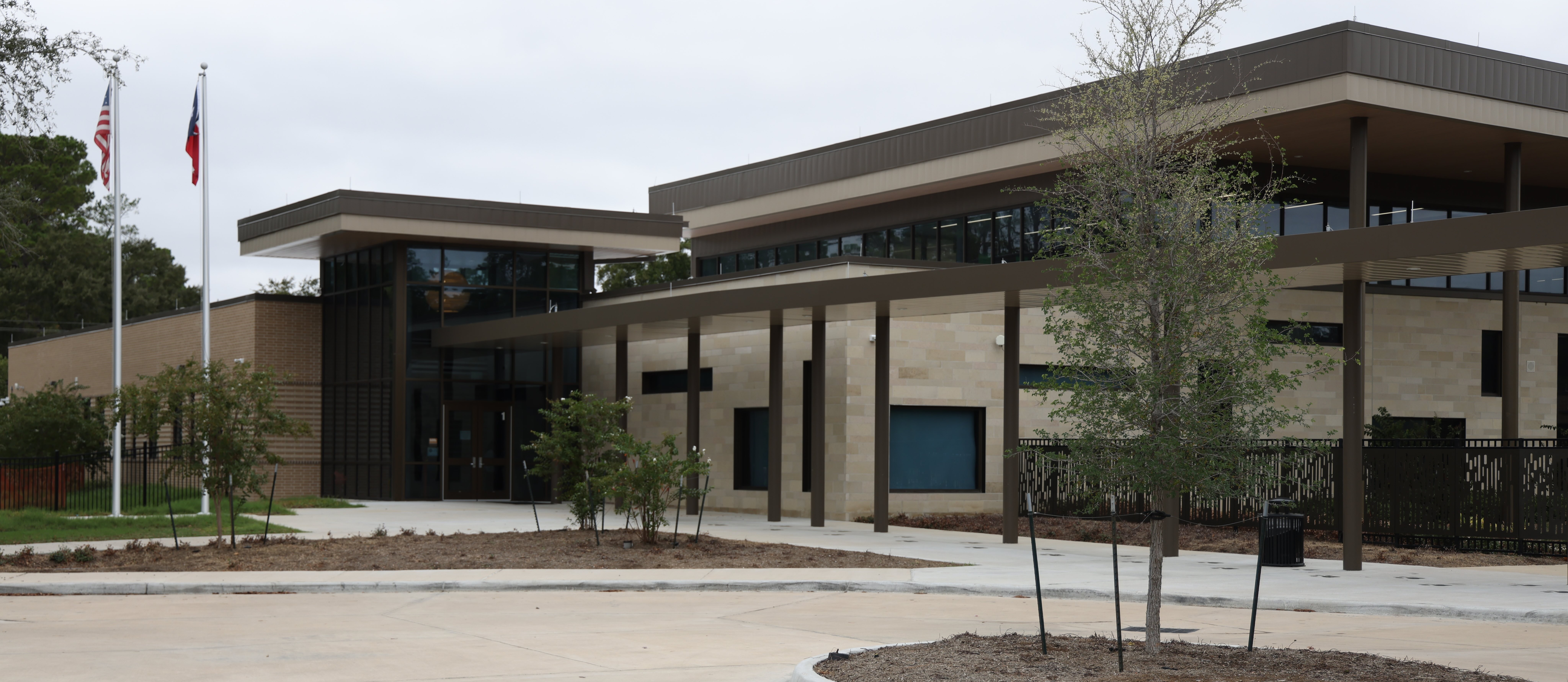 A modern building with a large entrance, flags, and greenery. The building has a flat roof and large windows.