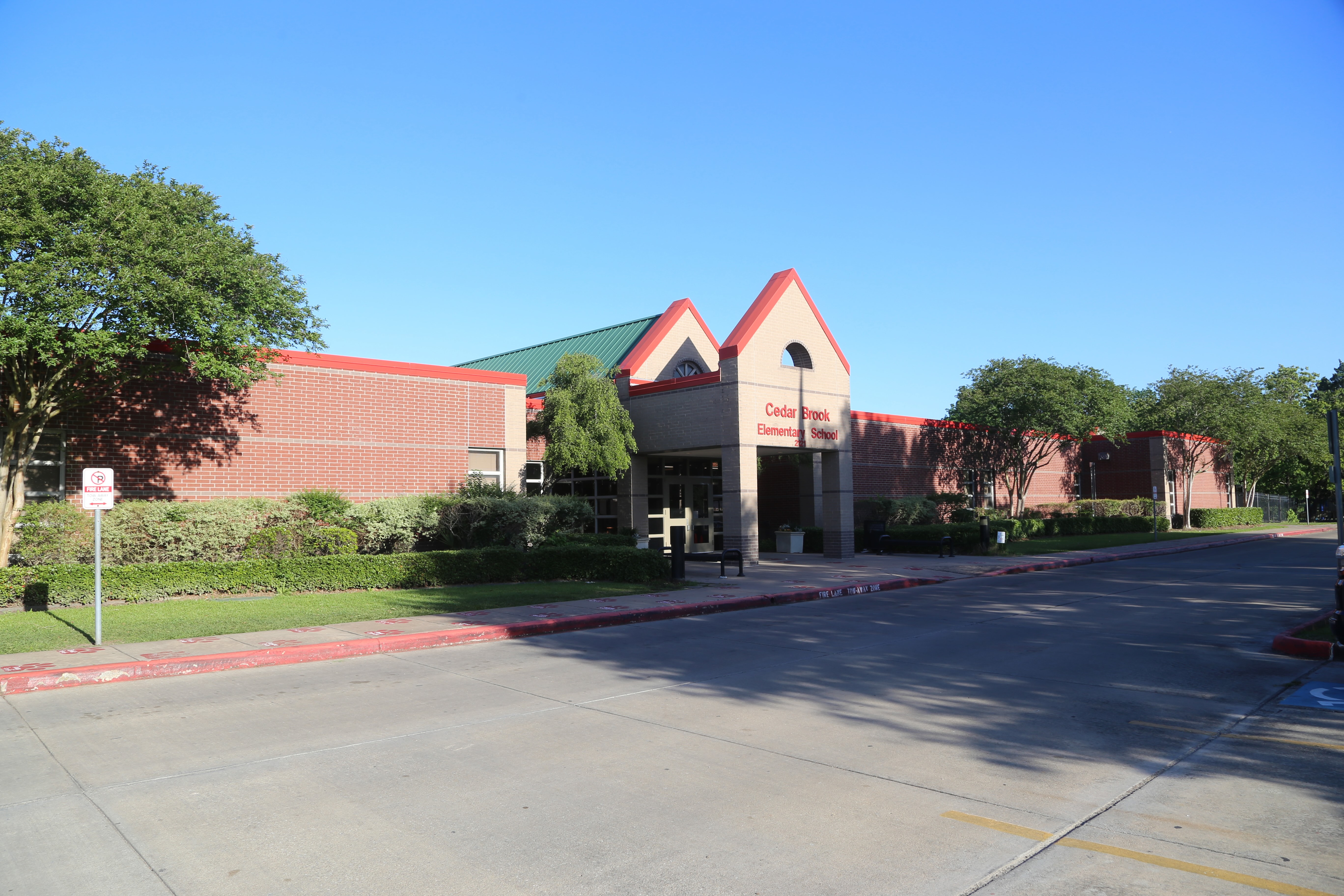 A large brick building with a green roof and red trim. Trees and bushes surround the building, and a parking lot is in front.