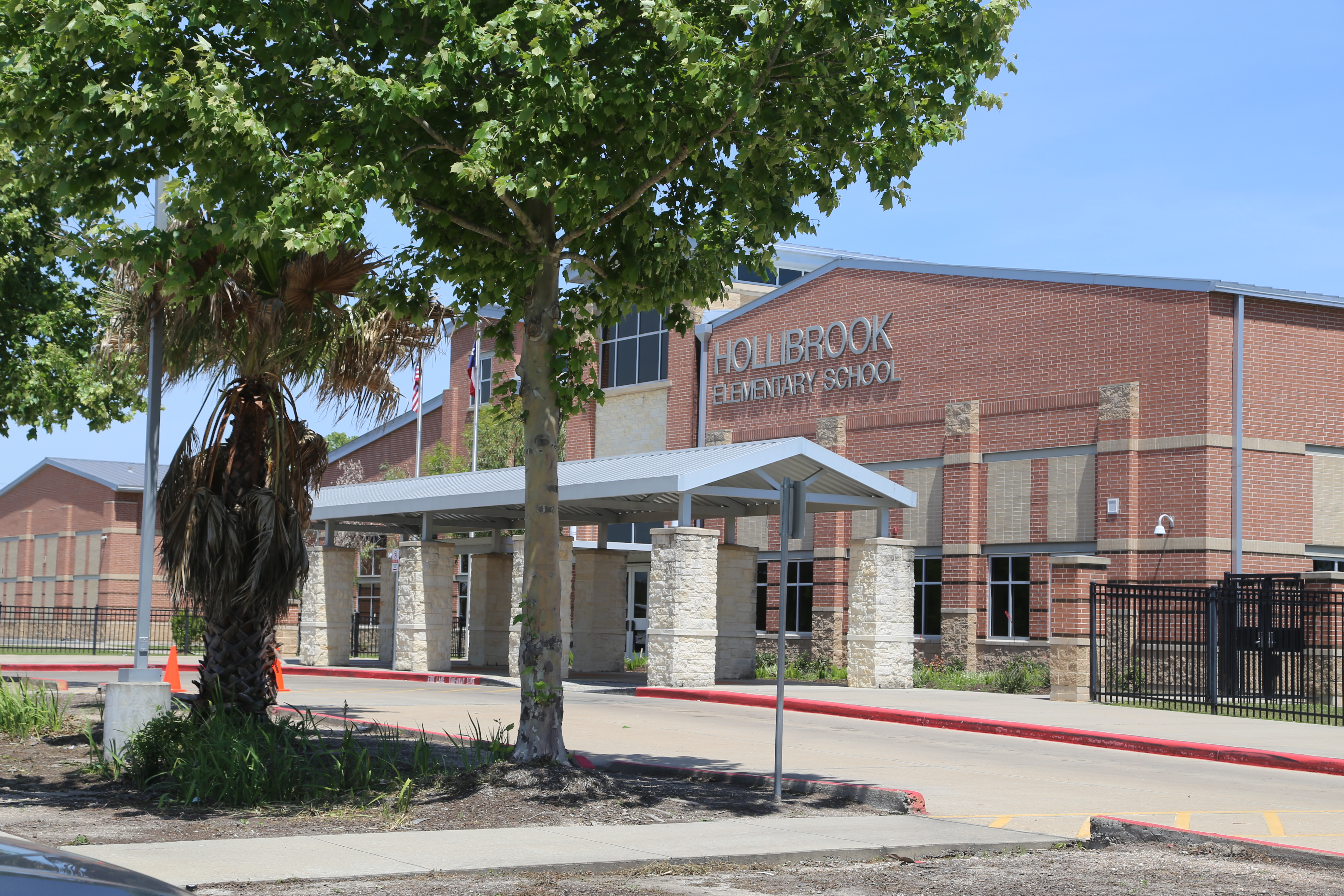 A school building with a brick exterior, large windows, and a covered entrance. A palm tree and a tree stand before it.