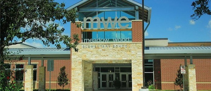 A building entrance with a glass canopy and brick pillars. "MWE" is written above the entrance. Trees and grass are in the foreground.