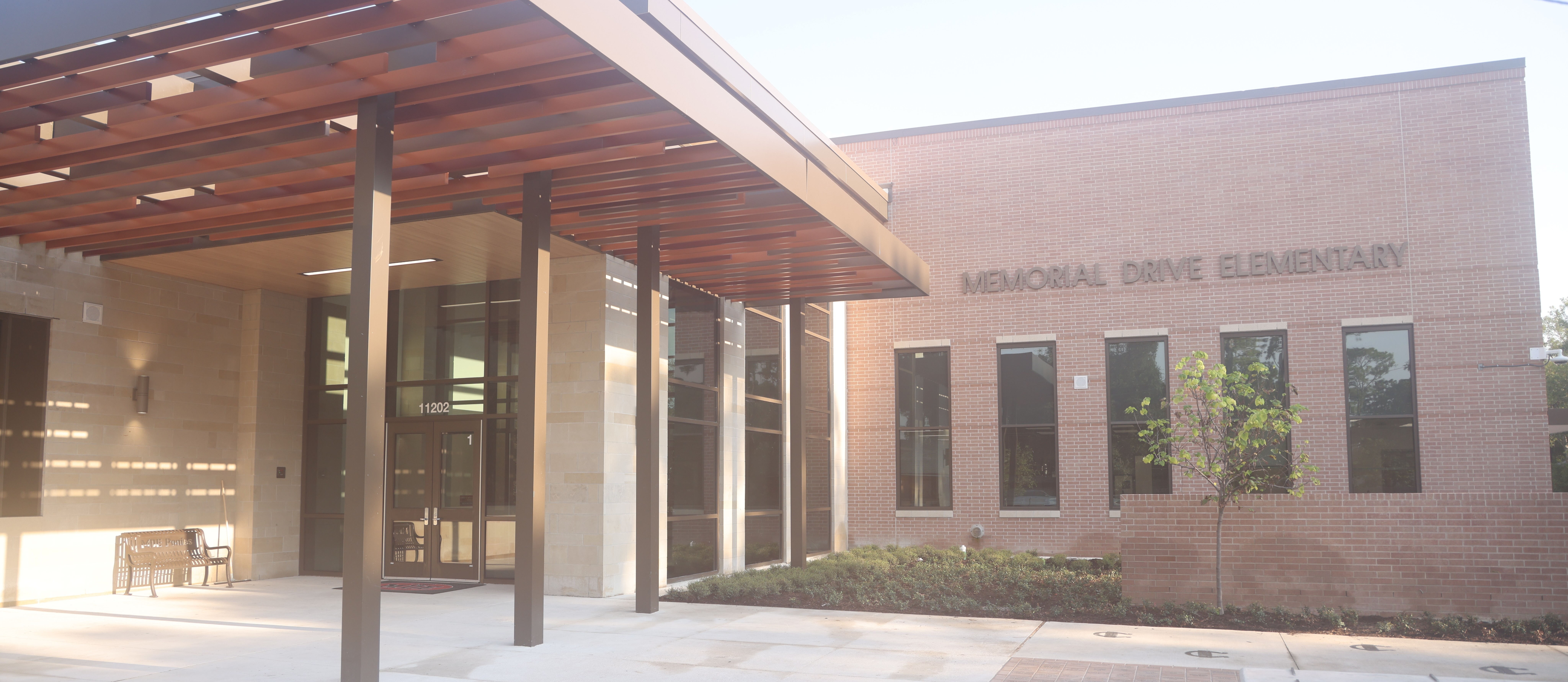 A modern building with glass windows, a covered entrance, and a bench, with the sign "Memorial Drive Elementary" on it.
