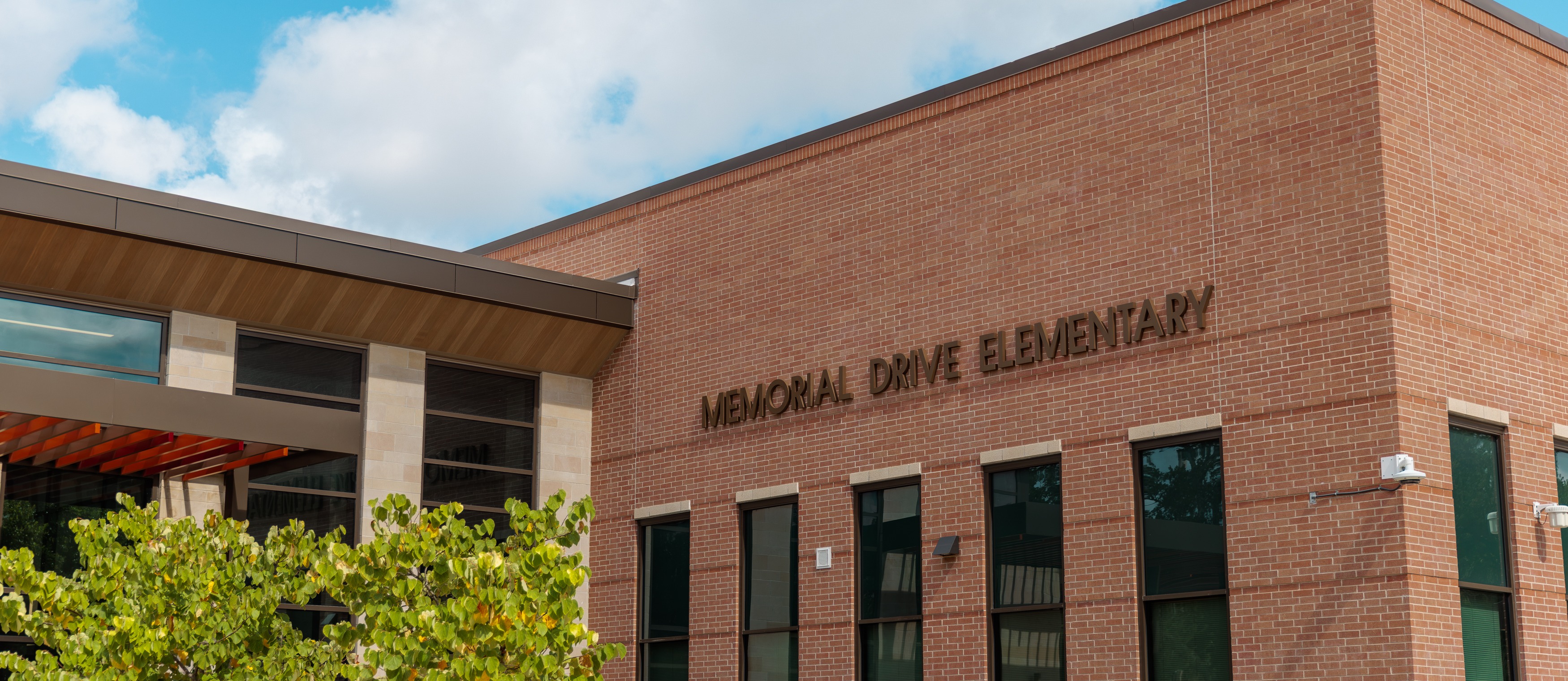 Front of a brick building with "Memorial Drive Elementary" text. Large windows, a tree, and a bicycle rack.