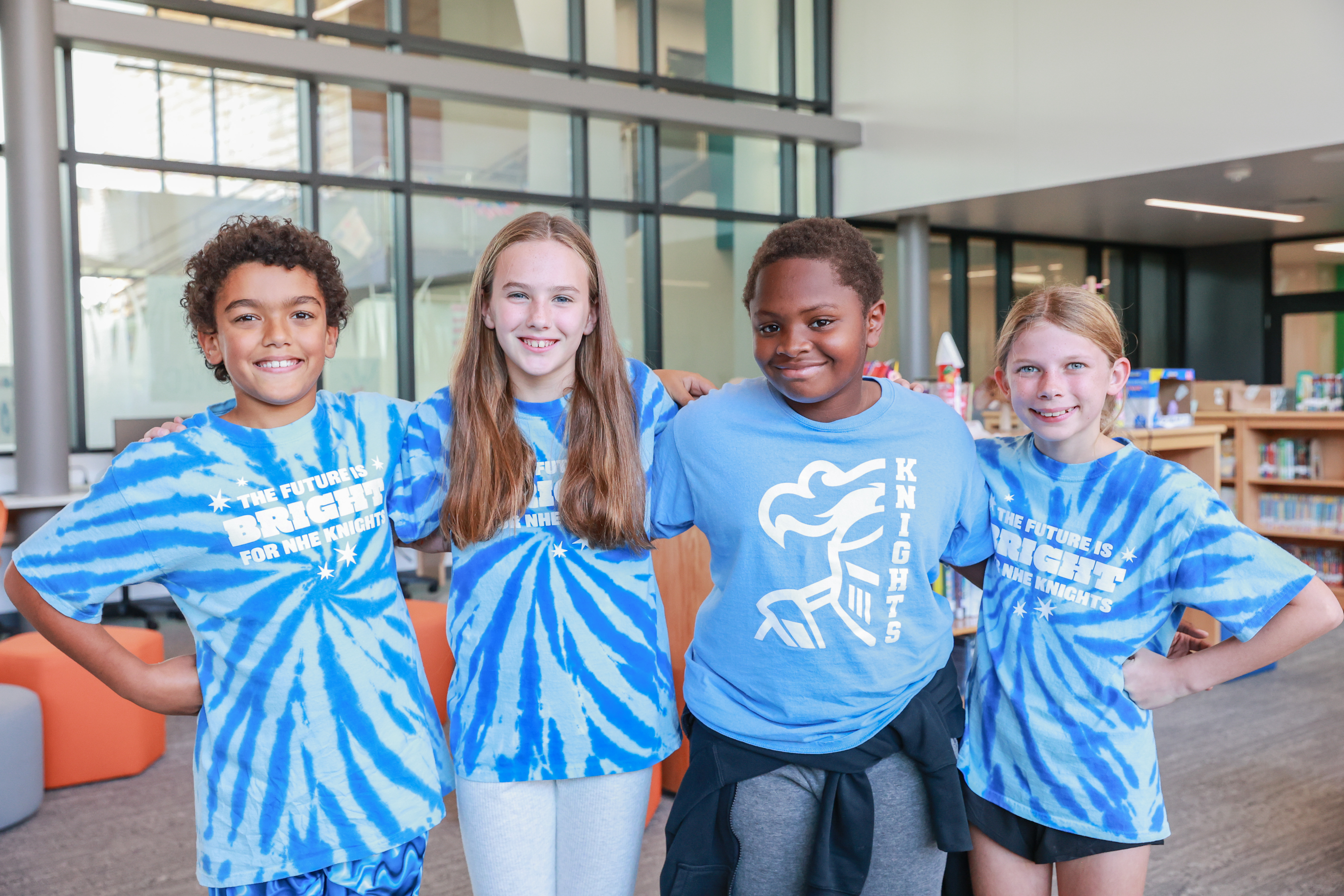 Four children wear blue tie-dye shirts with a logo. They stand close to each other in a room with shelves and glass windows.