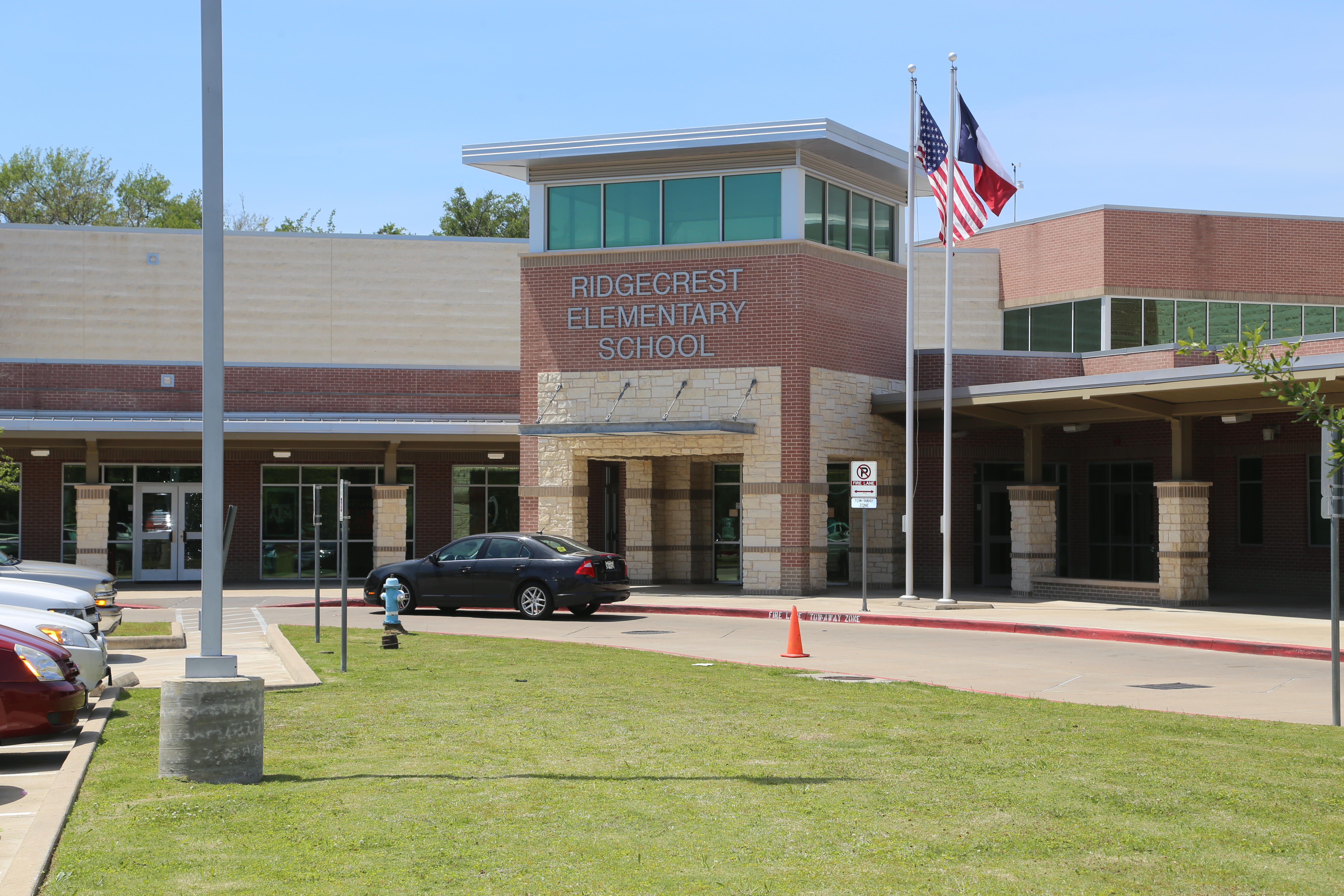 A school building with "RIDGECREST ELEMENTARY SCHOOL" above the entrance. Flags on poles in front. Cars parked outside.