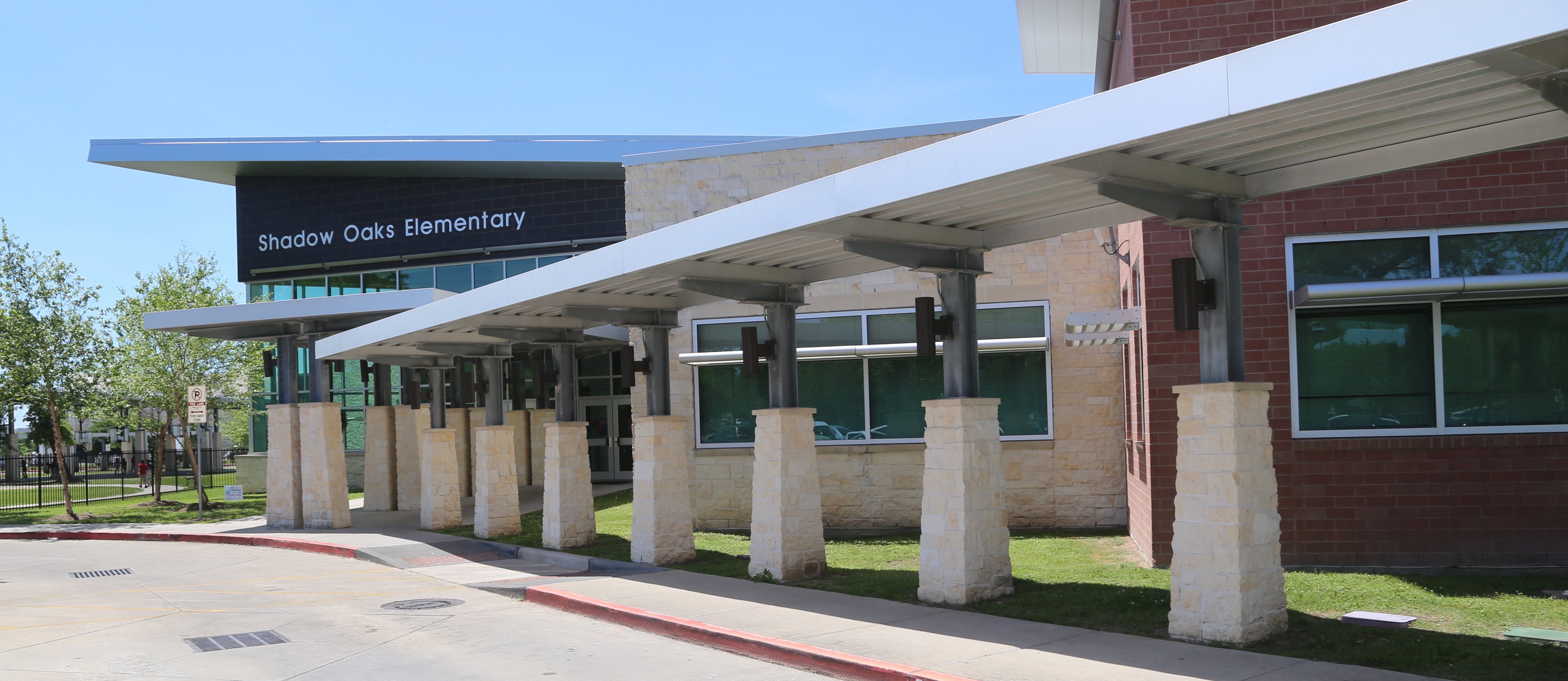 Exterior view of an elementary school building with a long awning. Trees, grass, and orange traffic cones are visible in the background.