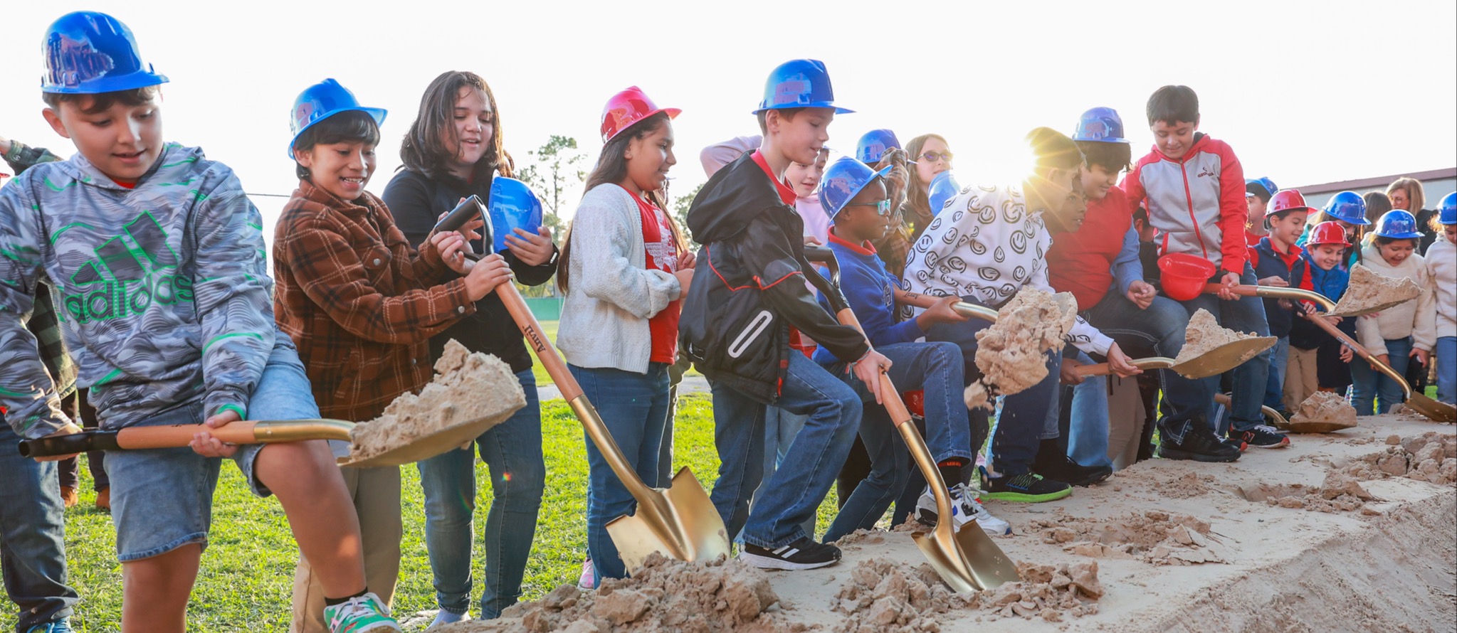 Children and adults in blue hats hold shovels, digging up sand. A line of people in the background.