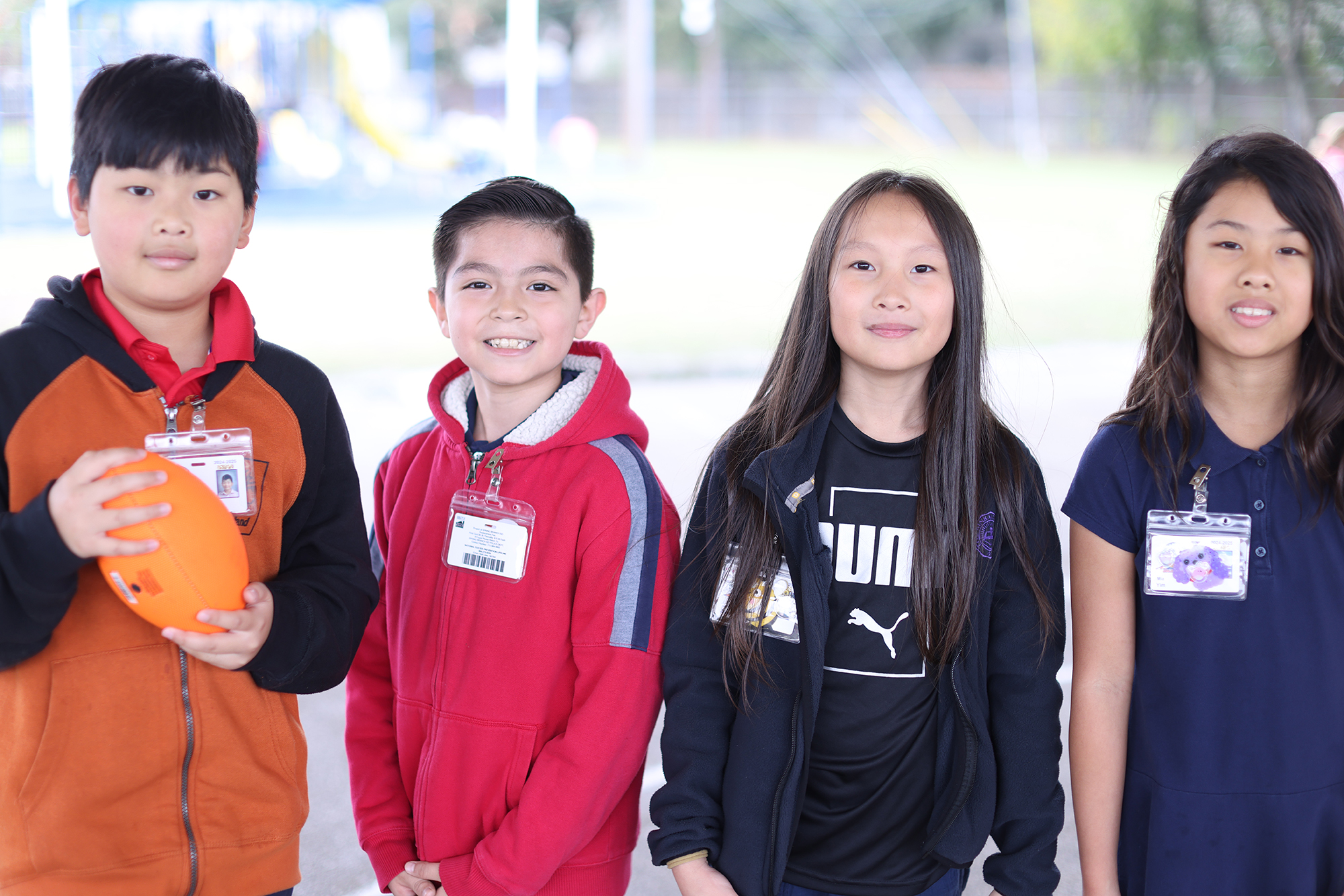 Four children in different colored tops stand outside. Two boys and two girls. One boy holds a football.