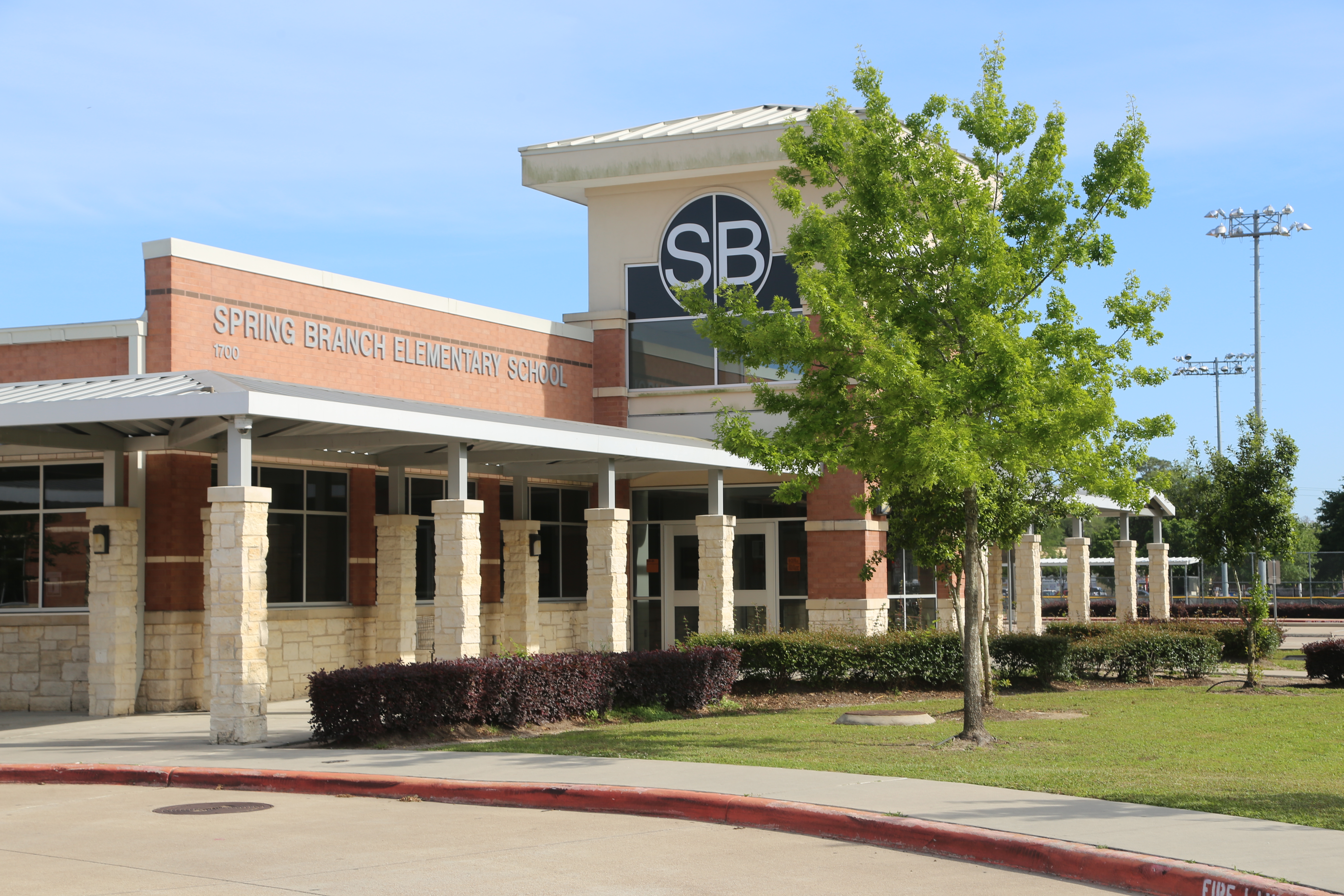 A school building with the letters "SB" on top. There is a tree and bushes in front.