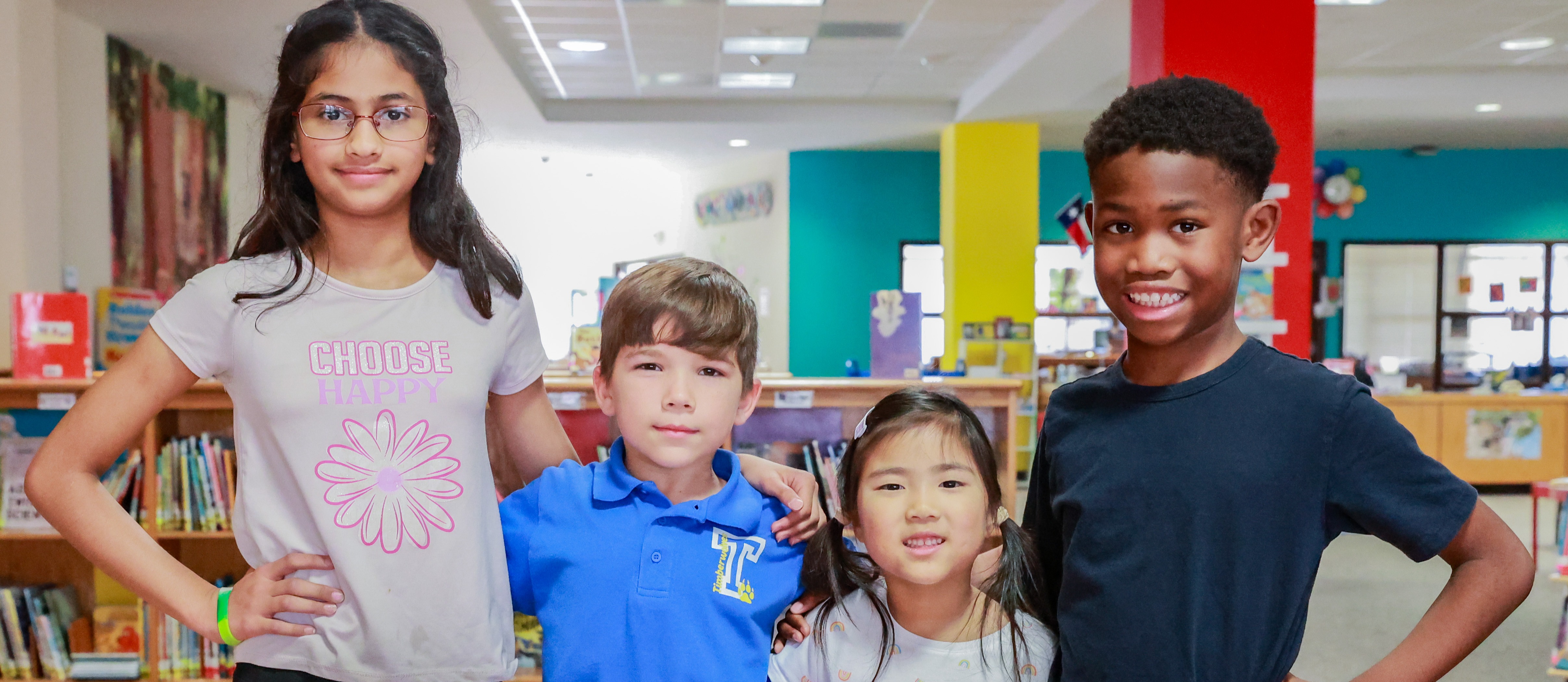 Four children pose for a photo in a brightly lit library with colorful walls and shelves filled with books.