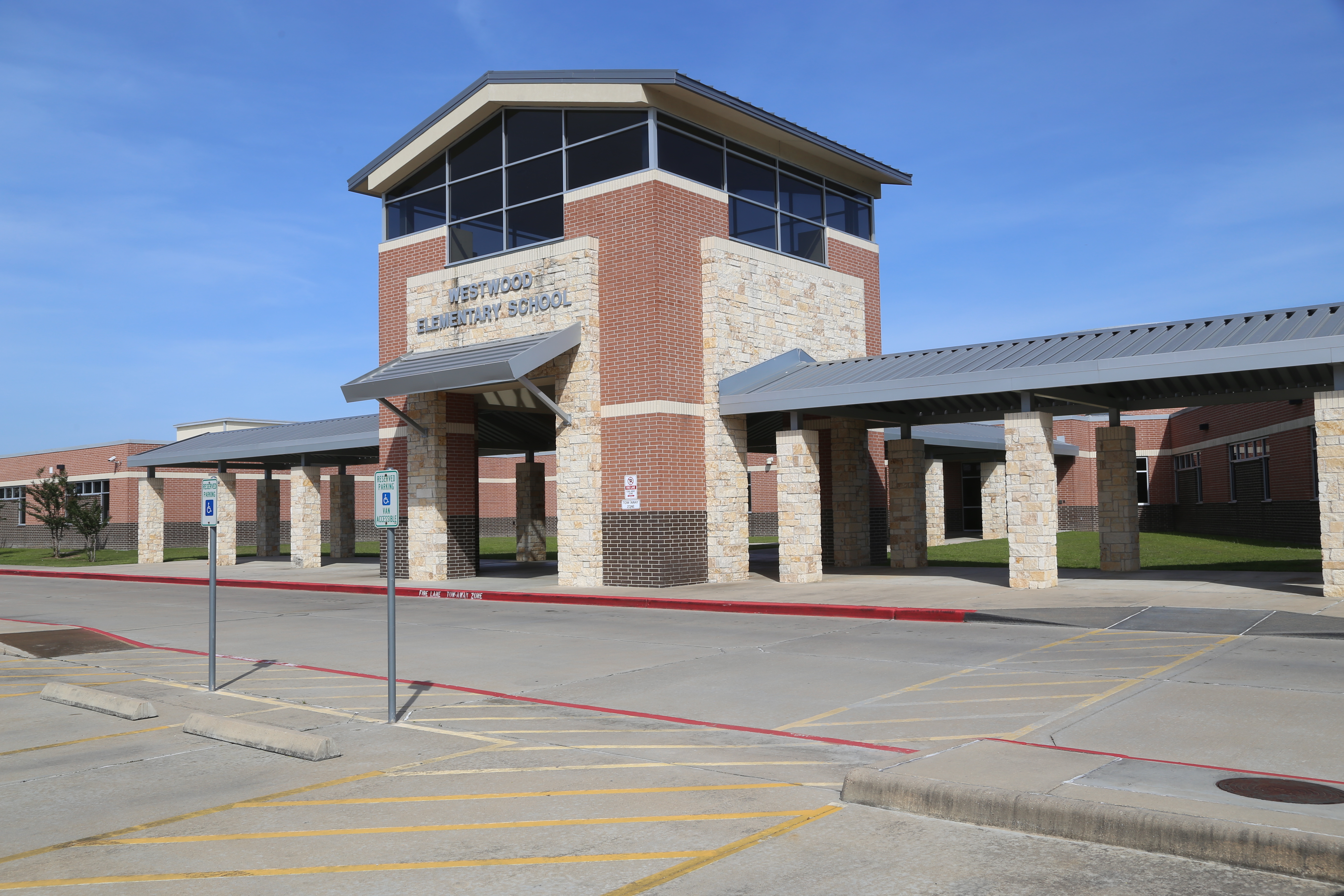 School building with brick walls, glass windows, and a gray roof. Empty parking lot in front.