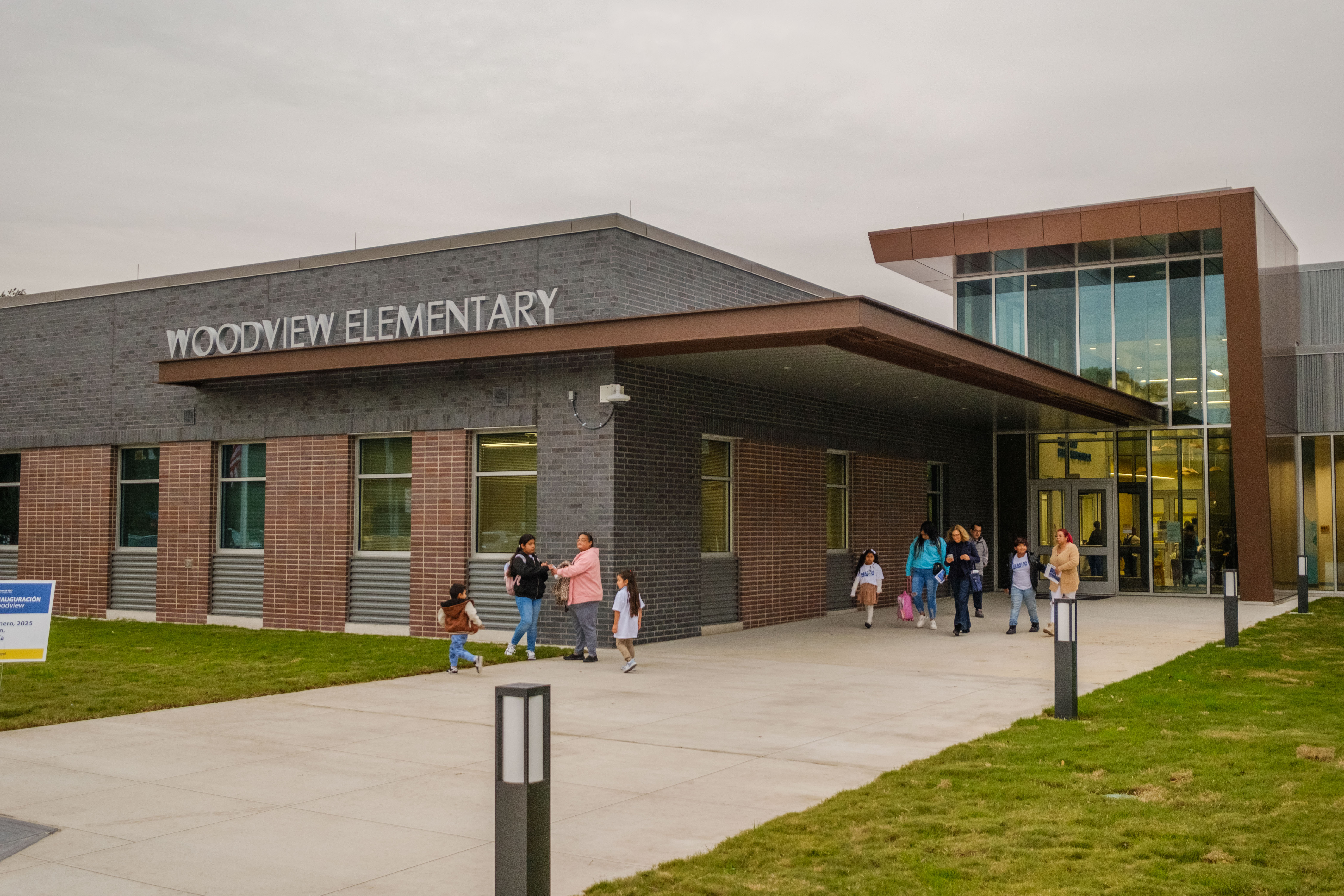 A modern school building with glass and brick exterior. People walk around in front of it.