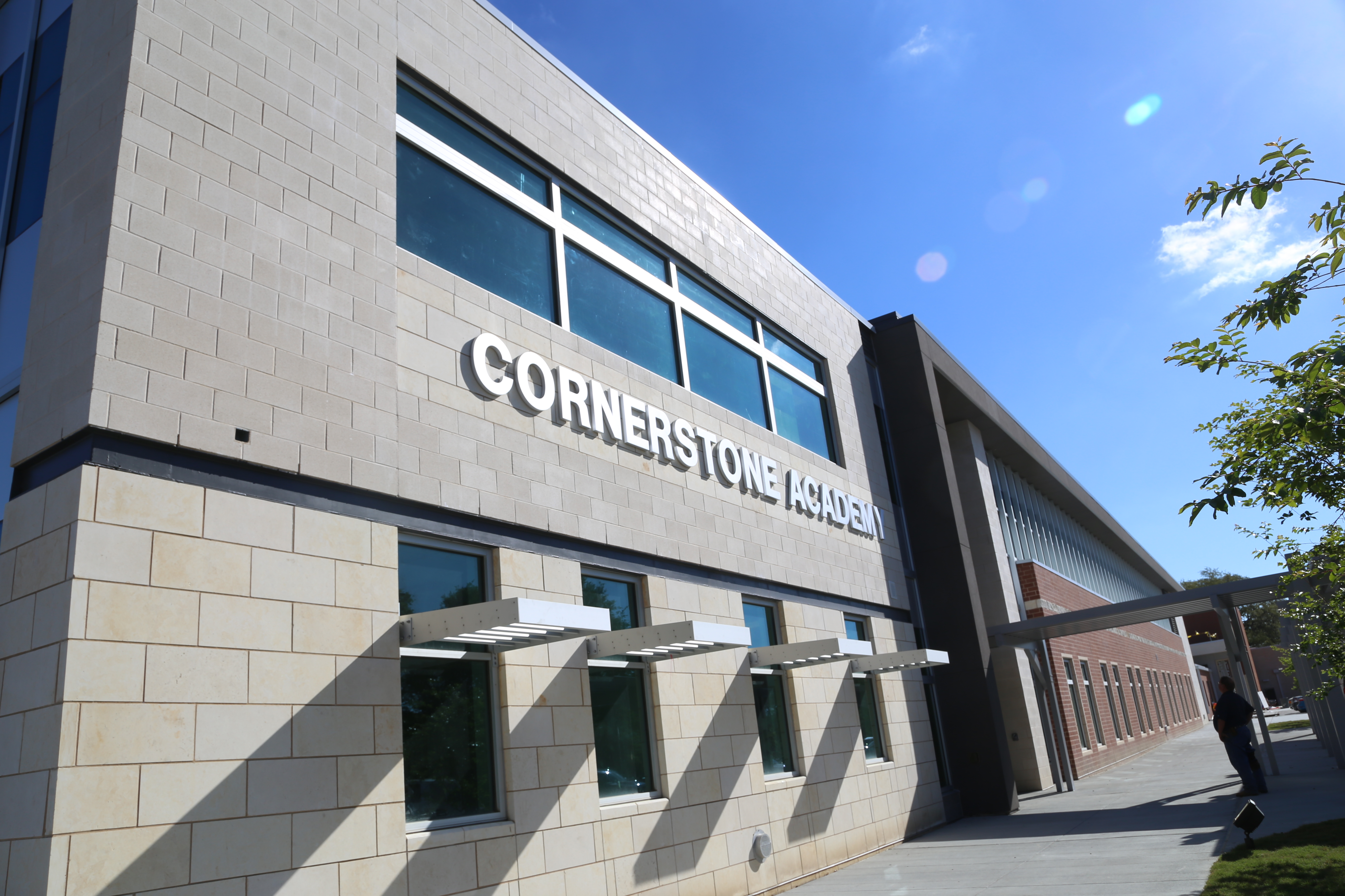 A building with "Cornerstone Academy" written on its side. It has many windows and a person walks past.