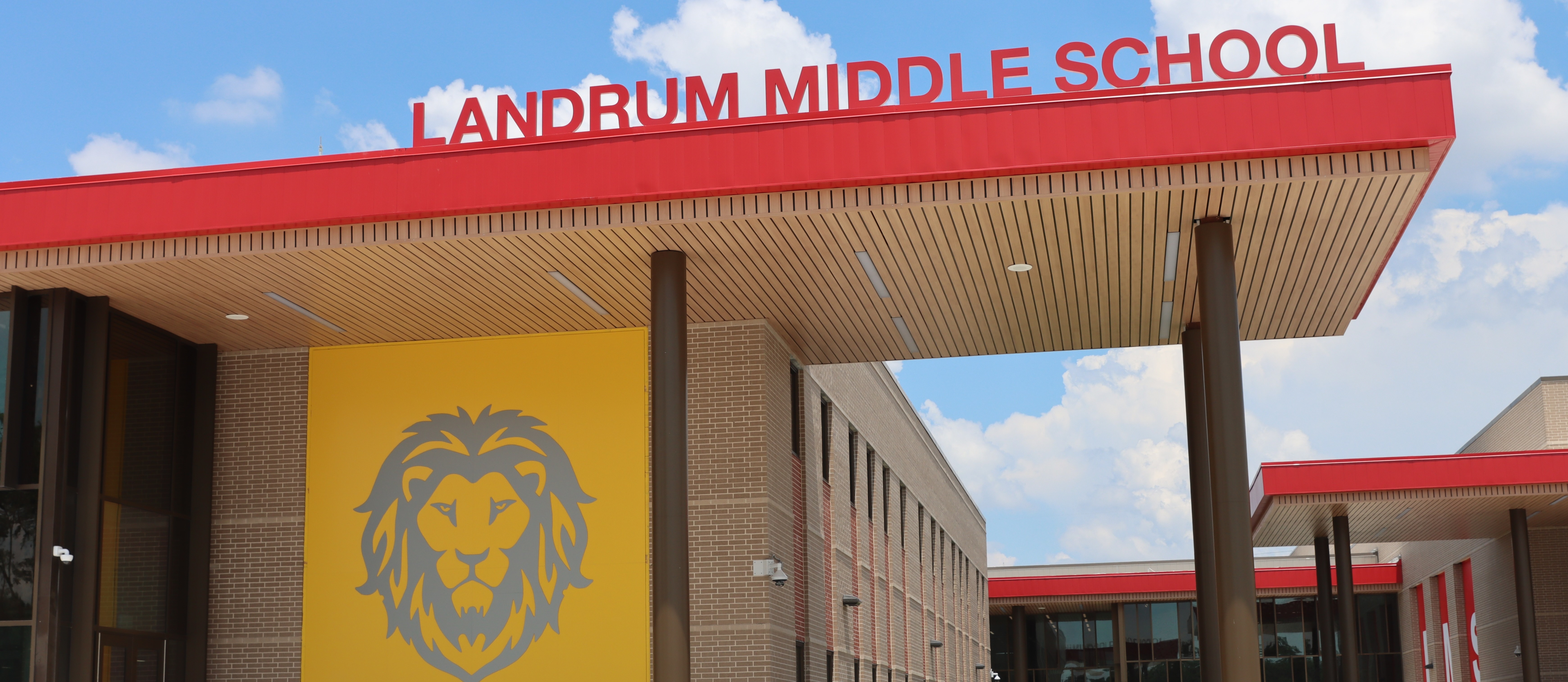 A building with a yellow wall featuring a lion logo. The sign reads "Landrum Middle School" over a red roof.