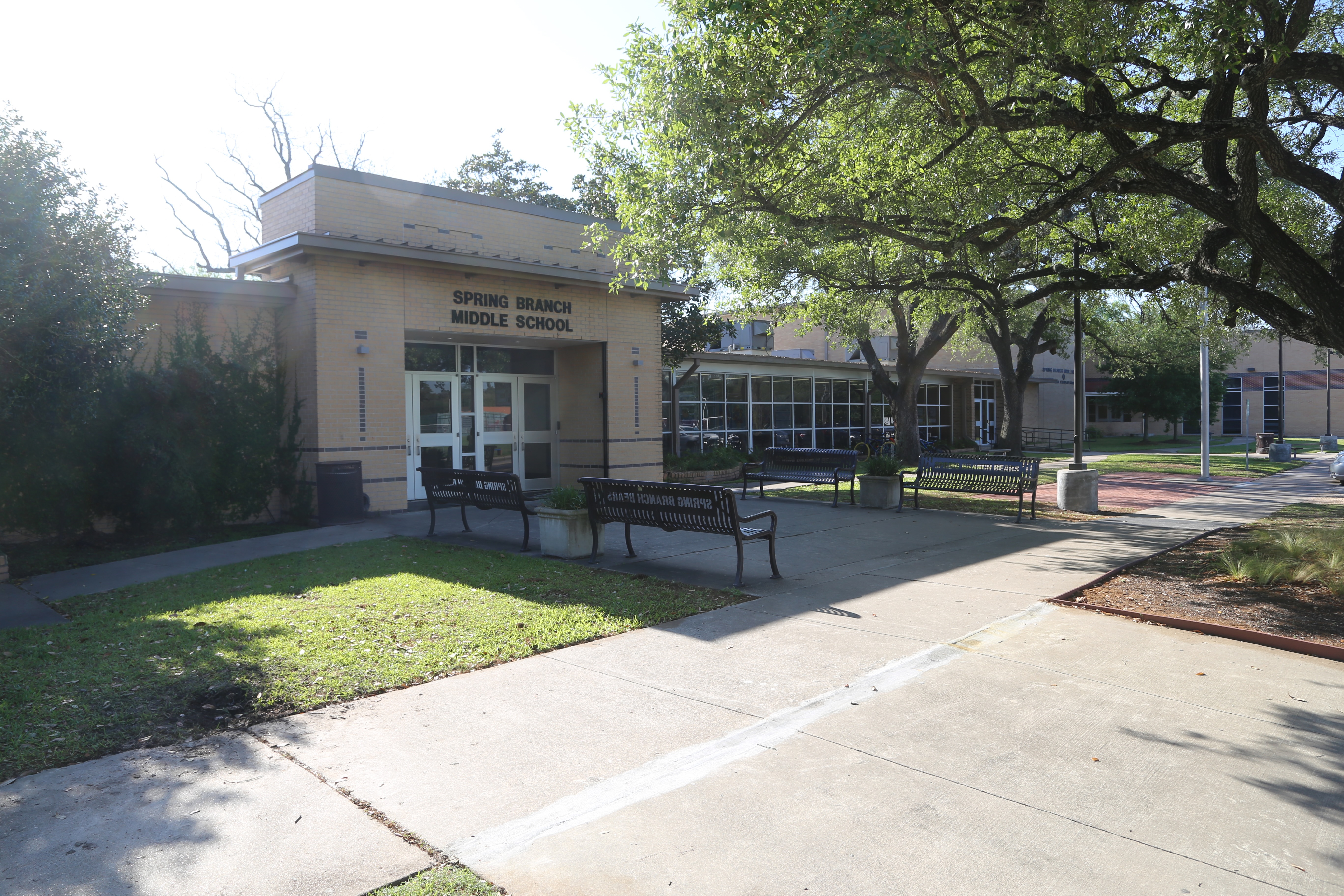 A building with a sign reading "Spring Branch Middle School" has benches on its front walkway. Trees and shrubs surround the area.