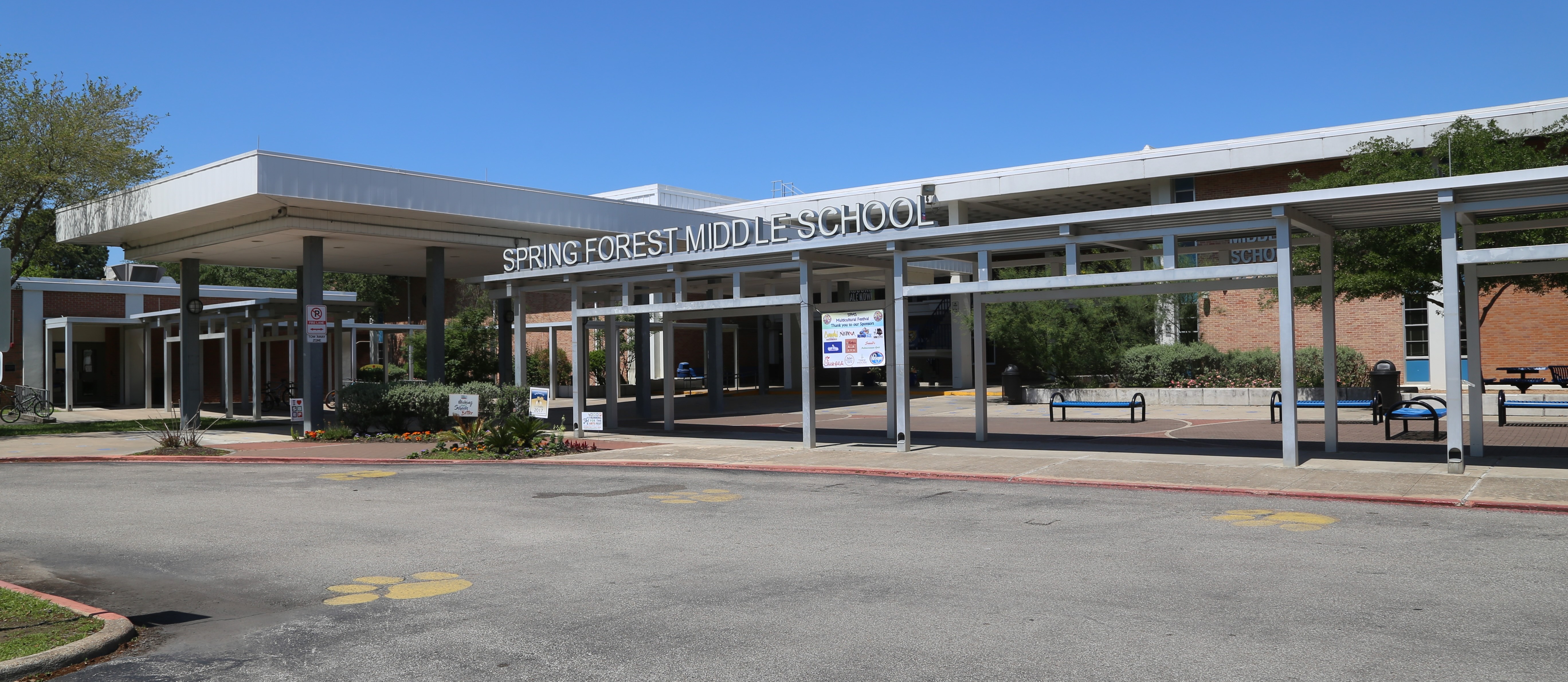 A school building with a white roof and metal supports. Trees and plants surround the area. The parking lot is empty.