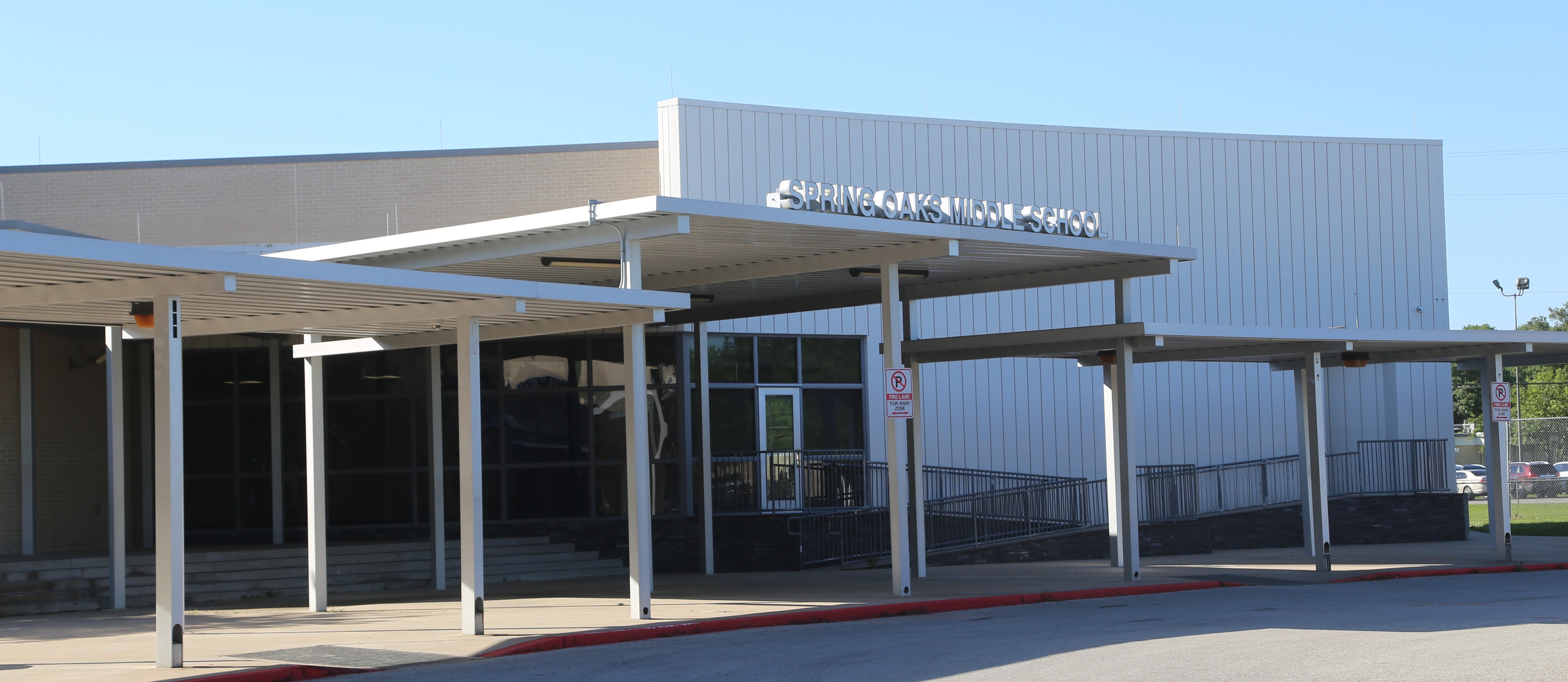 A modern building with a canopy and glass doors, under a blue sky with trees and parked cars in the distance.
