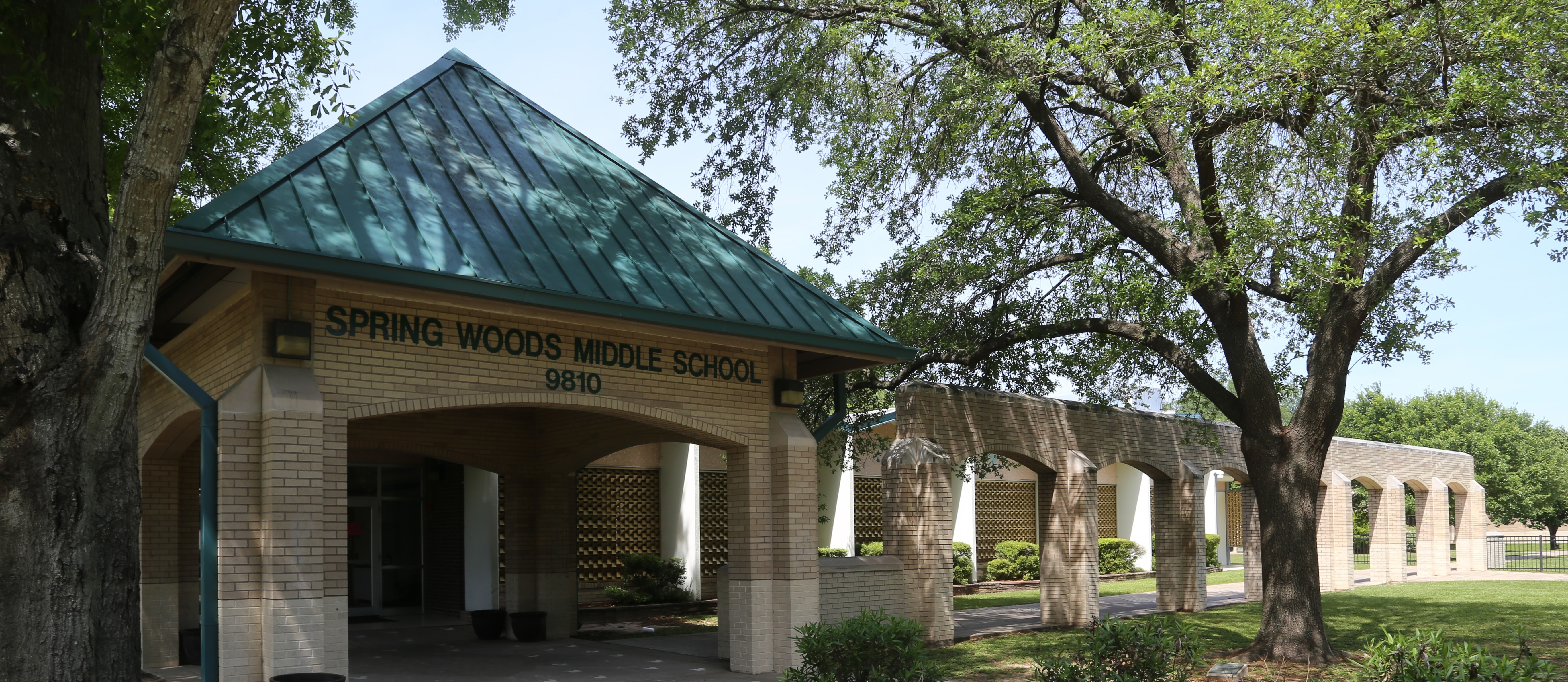 A building with a green roof has the text "Spring Woods Middle School" on it. Trees and shrubs are in front.