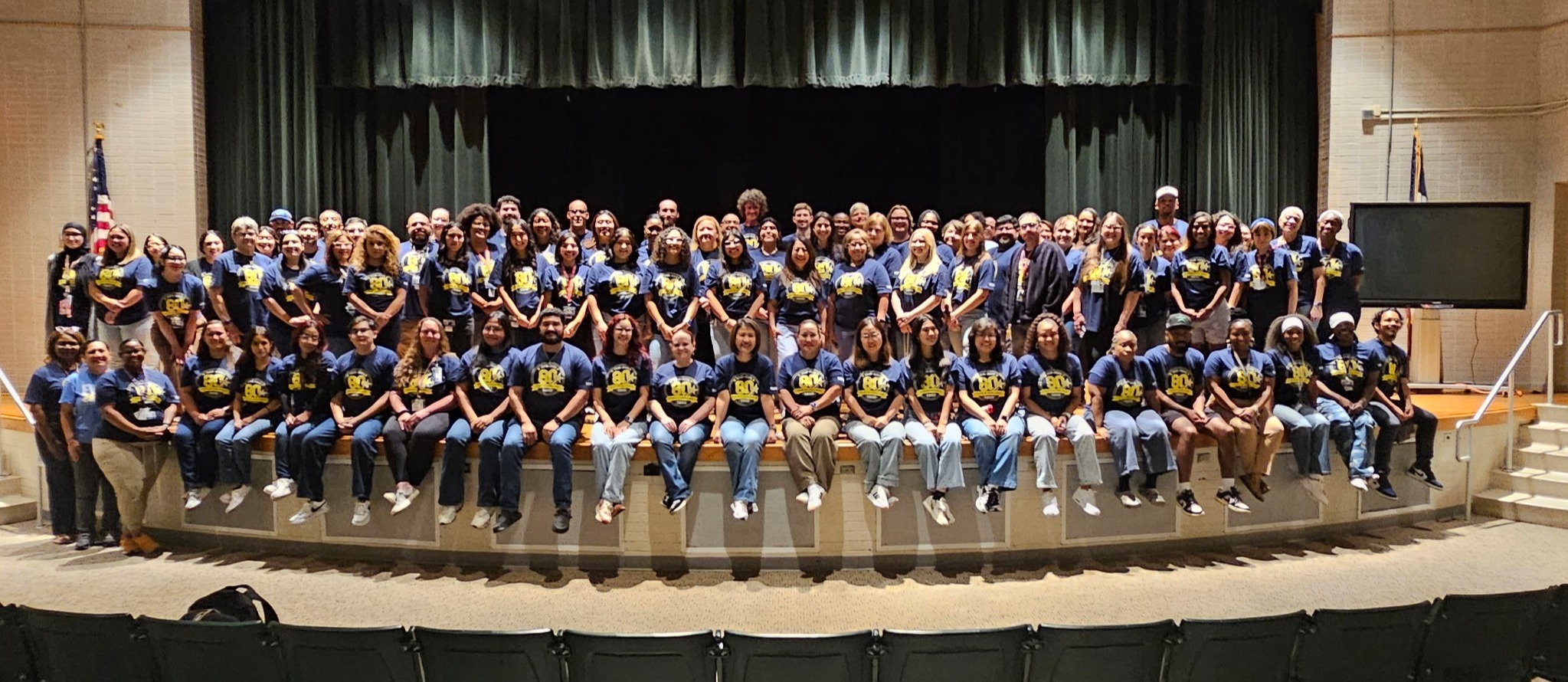 Large group in matching blue shirts with yellow and black patterns standing on a stage with a black curtain backdrop.