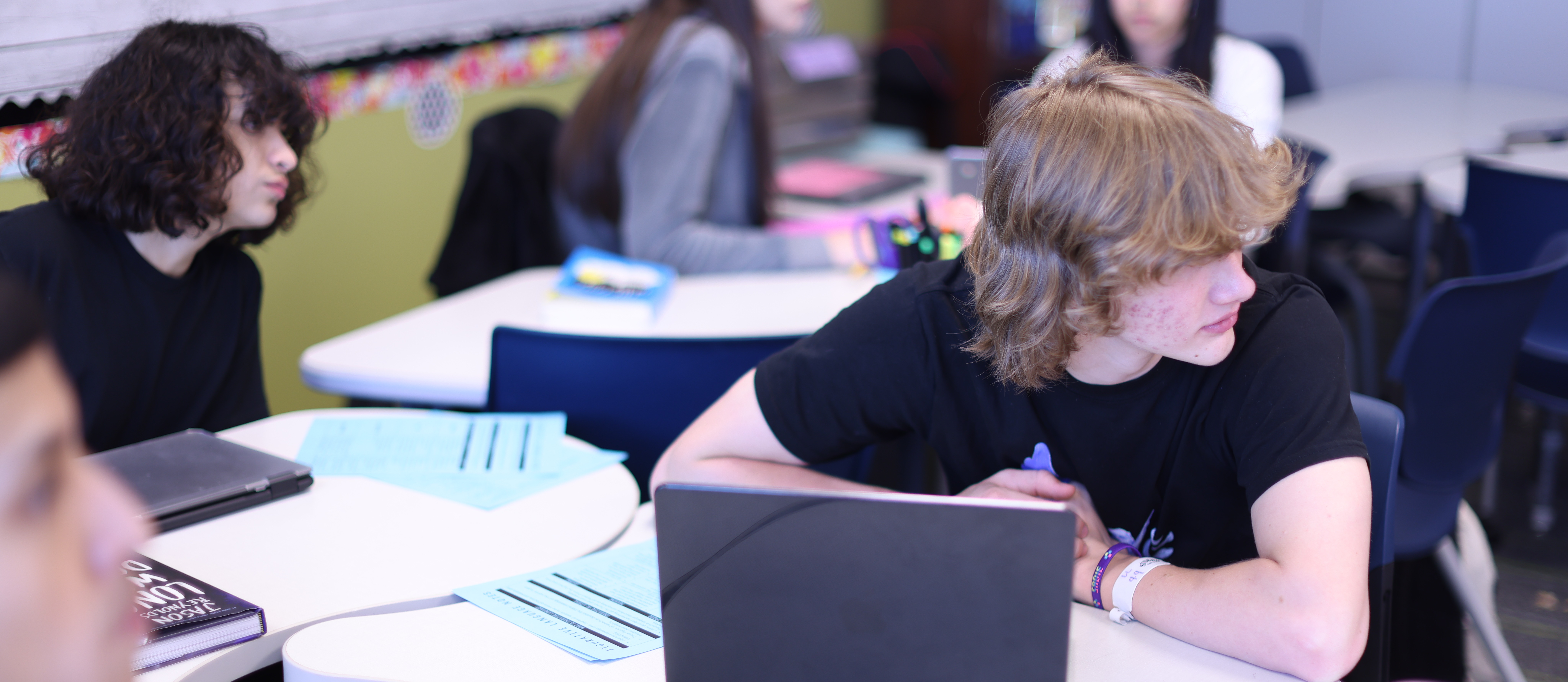 Four students sit at round tables in a classroom, focusing on laptops. Papers and pens are on the tables.
