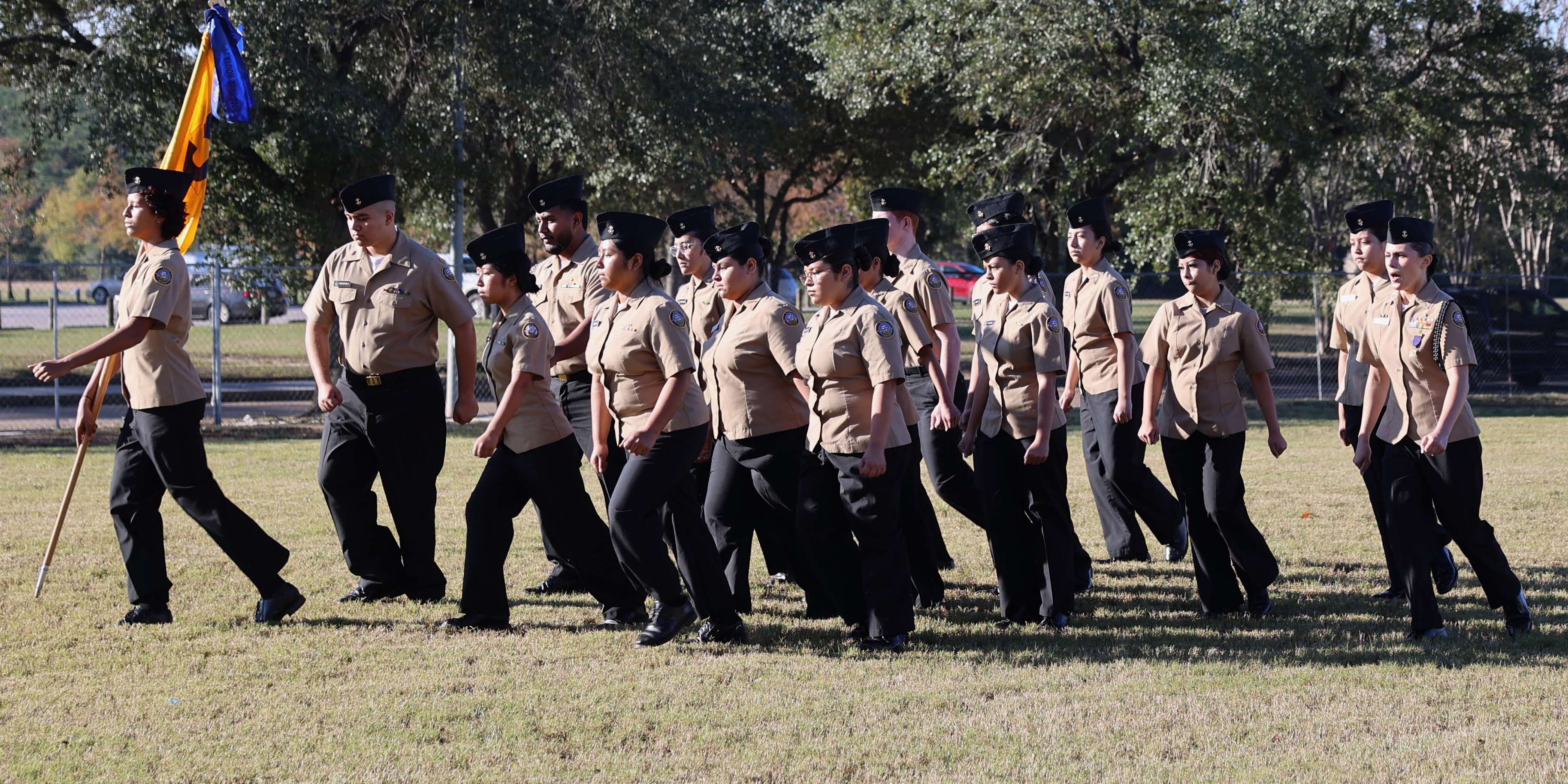 A group of uniformed individuals marches on a grassy field with trees in the background.