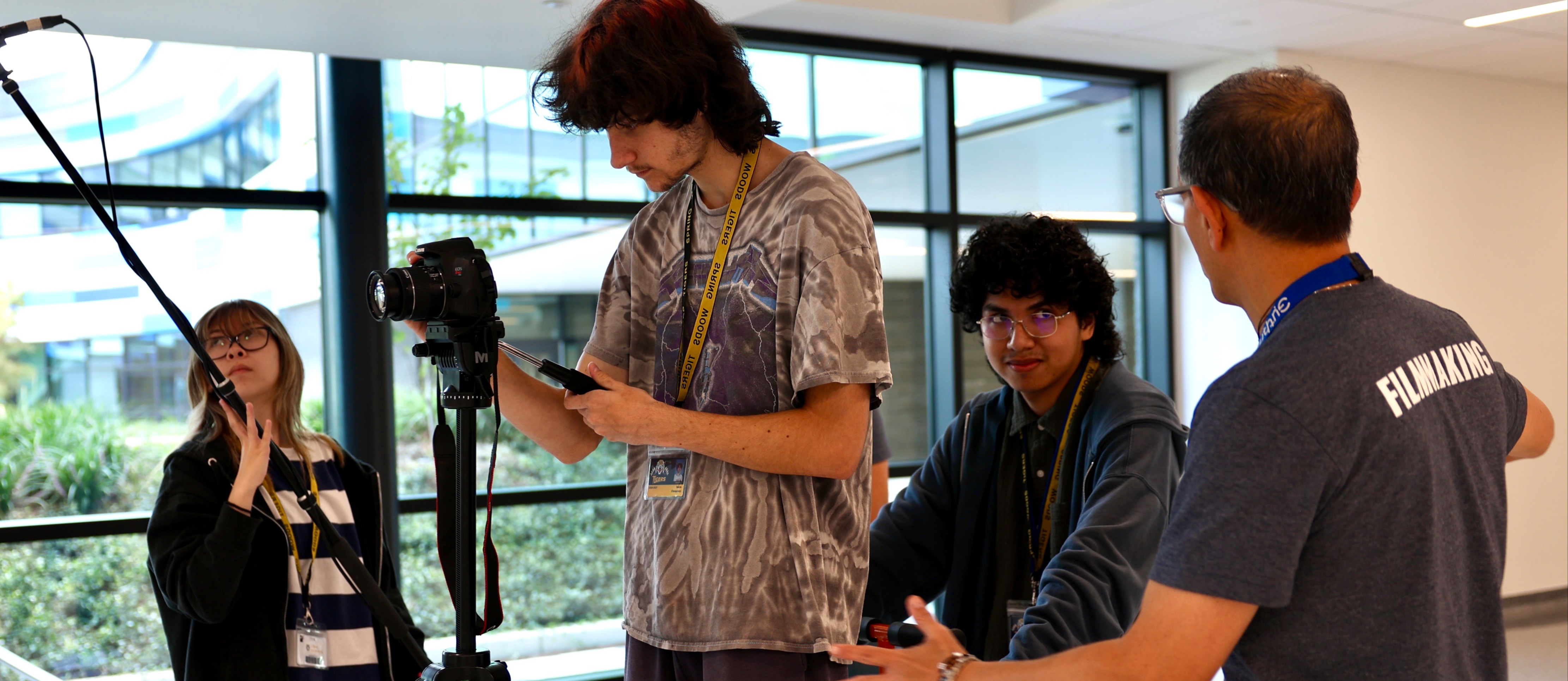 Four people are in a hallway, with one holding a camera and another gesturing. Background features glass windows.