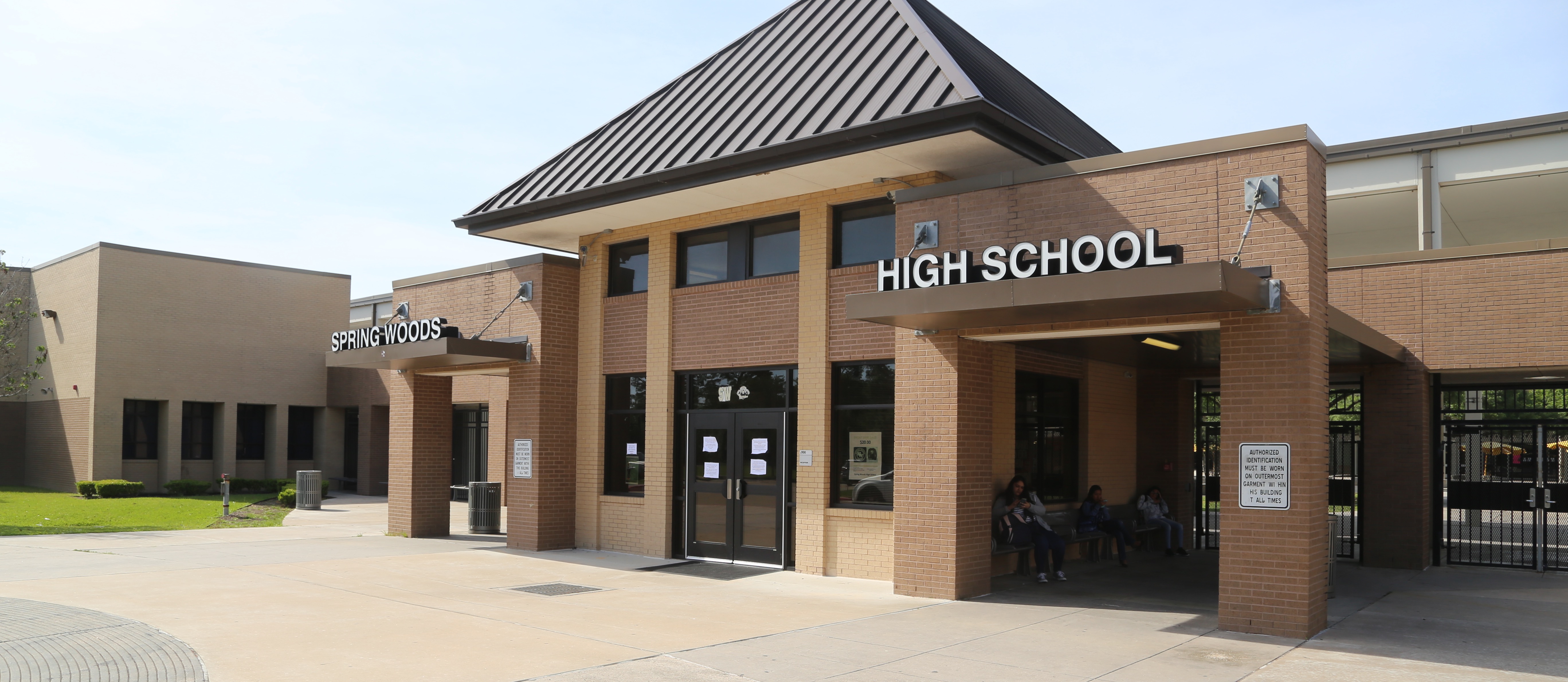 A high school building with a gray roof, brick walls, and large windows. A sign reads "HIGH SCHOOL".