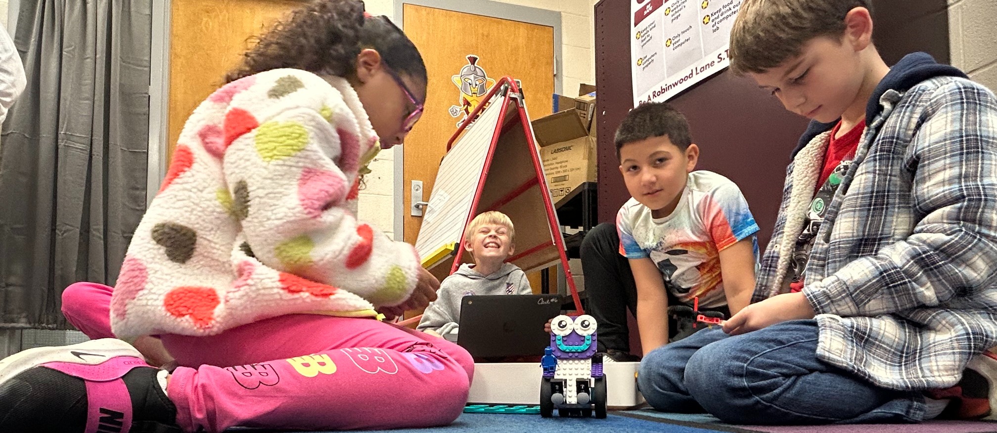 four students on rug in classroom working with Legos and coding