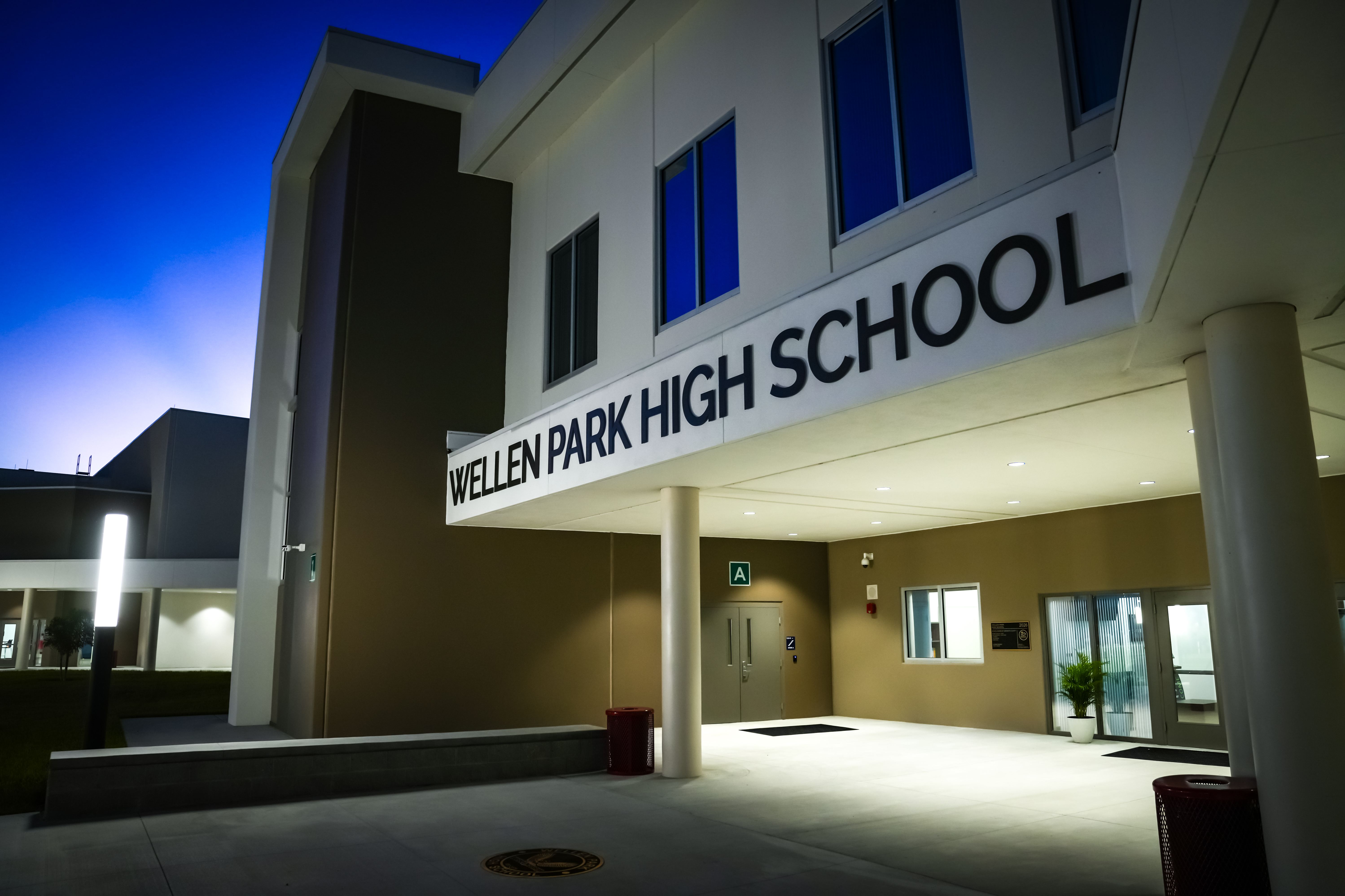 A school entrance at night, "Wellen Park High School" in bold letters above the entrance. The area is well-lit, and there are trash bins on either side.