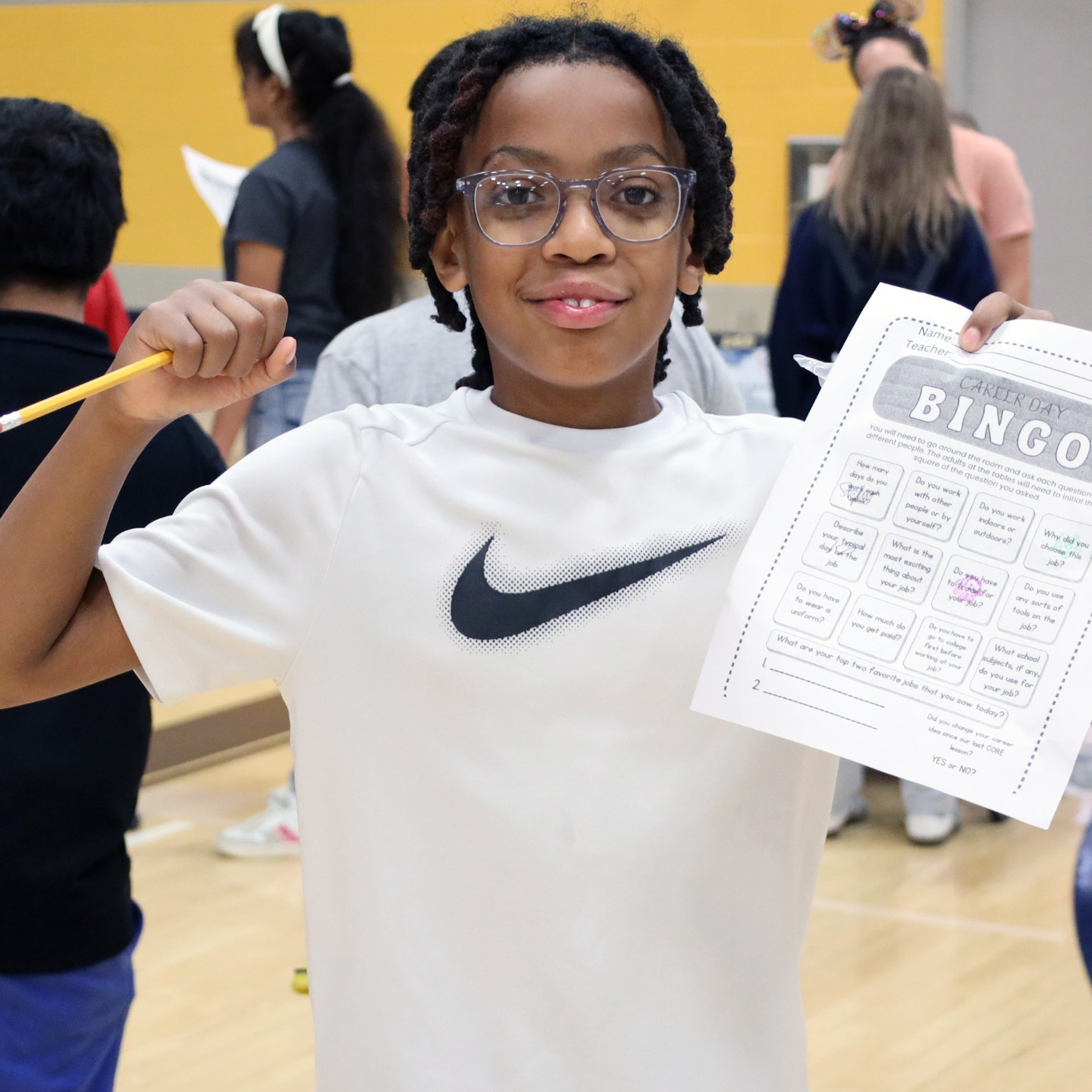 A young person in a white T-shirt and glasses holds up a bingo card with pencils.