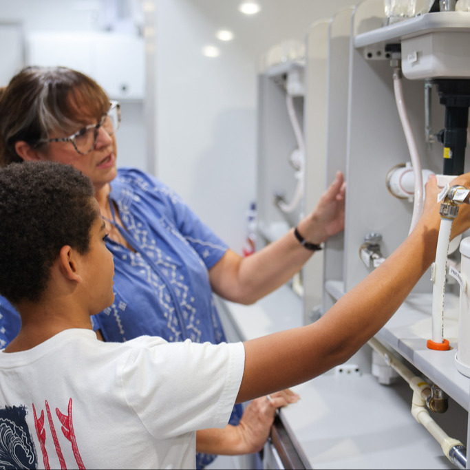 A woman with glasses and a watch assists a child in a white shirt in fixing a machine in a lab.